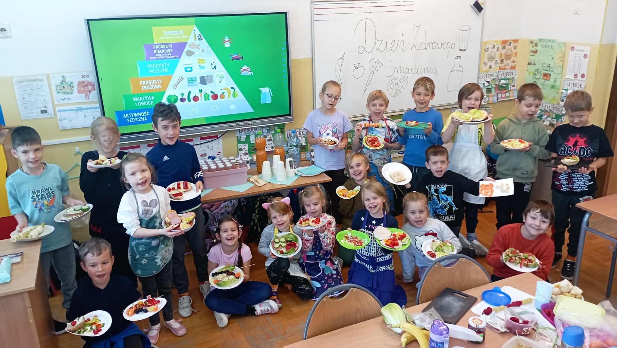 W sali lekcyjnej przed tablicą multimedialną wyświetlającą piramidę zdrowego żywienia ustawieni w dwa rzędy uczniowie klasy 1c z talerzykami w dłoniach. Na talerzykach kolorowe kanapki, sałatki i szaszłyki. Obok uczniów stolik z butelkami wyciskanych soków.