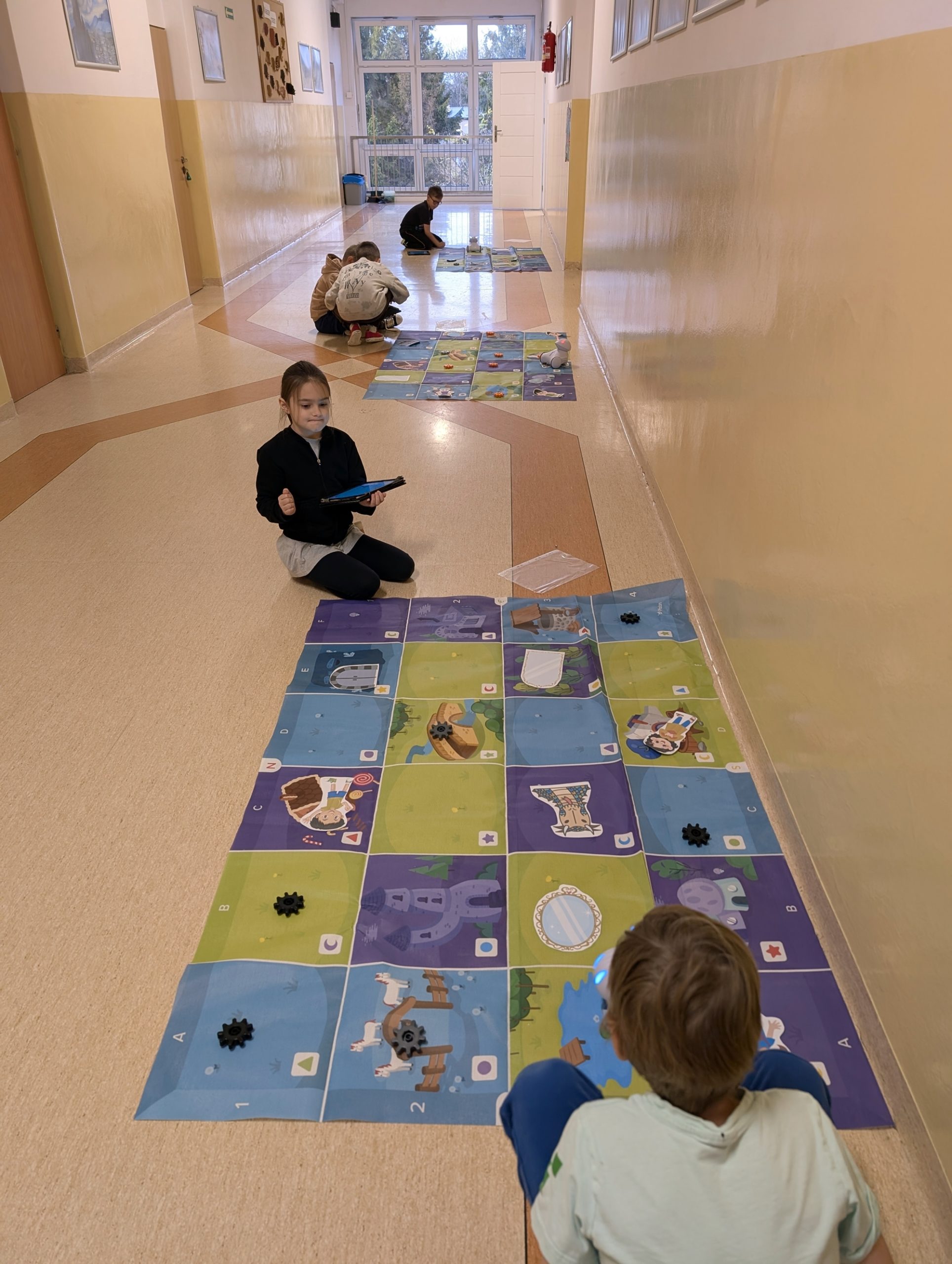 Dzieci siedzą na podłodze szkolnego korytarza i pracują z matami do programowania oraz małymi robotami. Na matach znajdują się ilustracje i elementy przypominające przeszkody. Jedno z dzieci trzyma tablet.