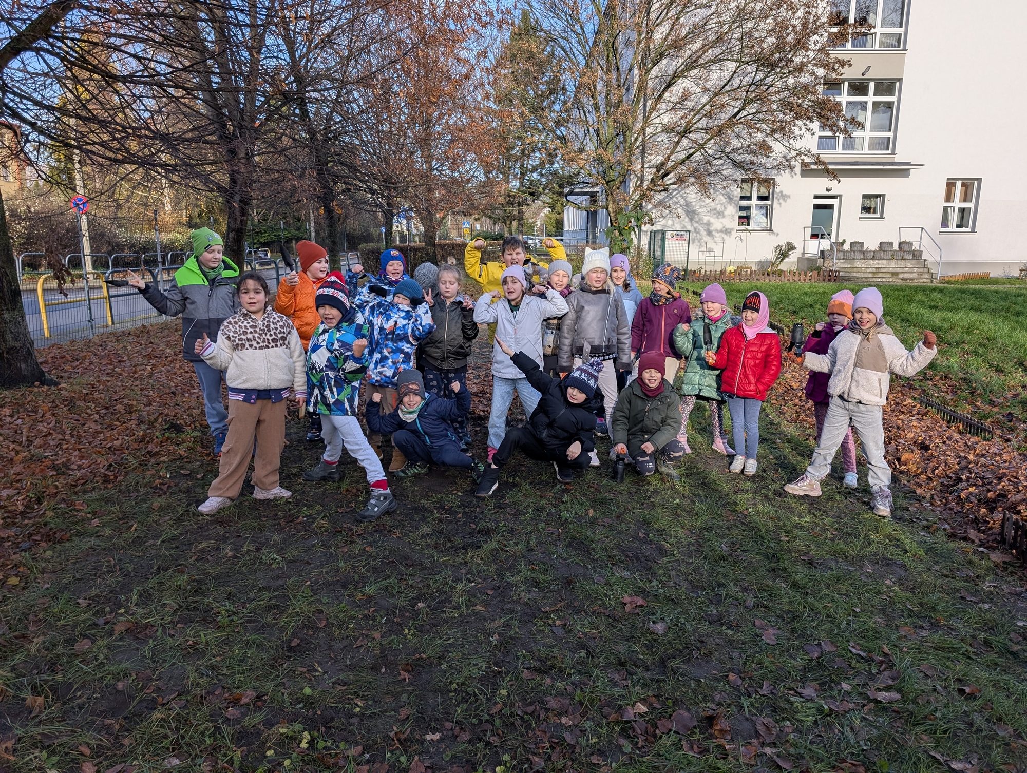 Grupa dzieci stojąca na trawniku w jesiennej scenerii, ubrane w kolorowe kurtki i czapki. Dzieci pozują radośnie do zdjęcia, a za nimi widać drzewa bez liści oraz budynek mieszkalny.