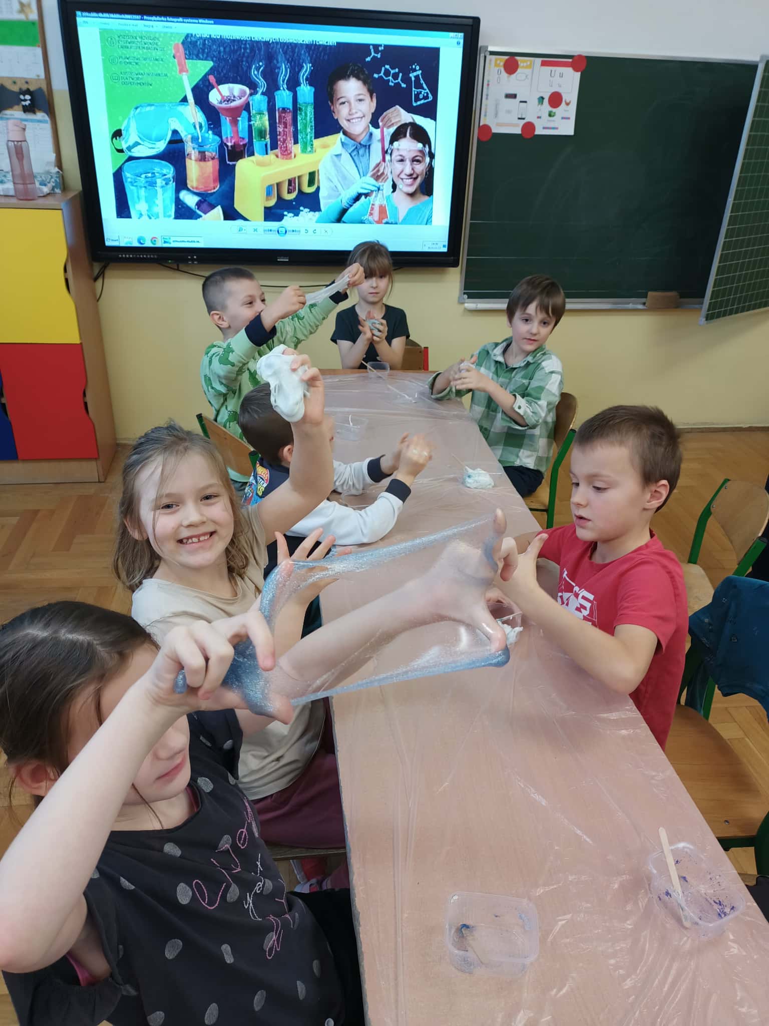 Uczniowie przy szkolnym stole rozciągają brokatowy slime podczas zajęć w klasie; w tle tablica multimedialna z grafiką doświadczeń.