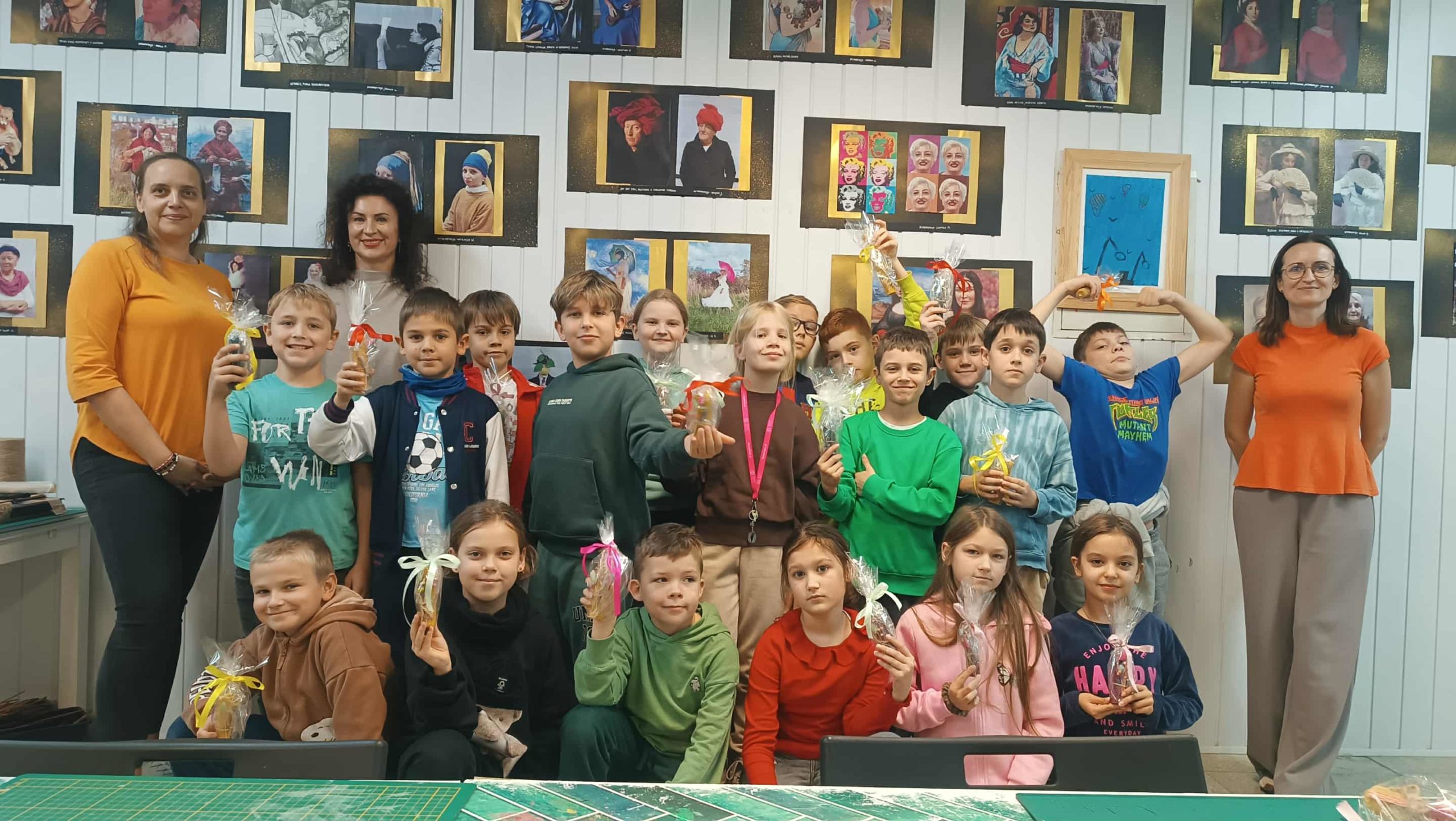 Grupowe zdjęcie całej klasy wraz z opiekunkami. Dzieci stoją i siedzą, trzymając zapakowane w folię własnoręcznie wykonane świeczki z wosku pszczelego. W tle widoczne są kolorowe prace artystyczne zawieszone na ścianie.