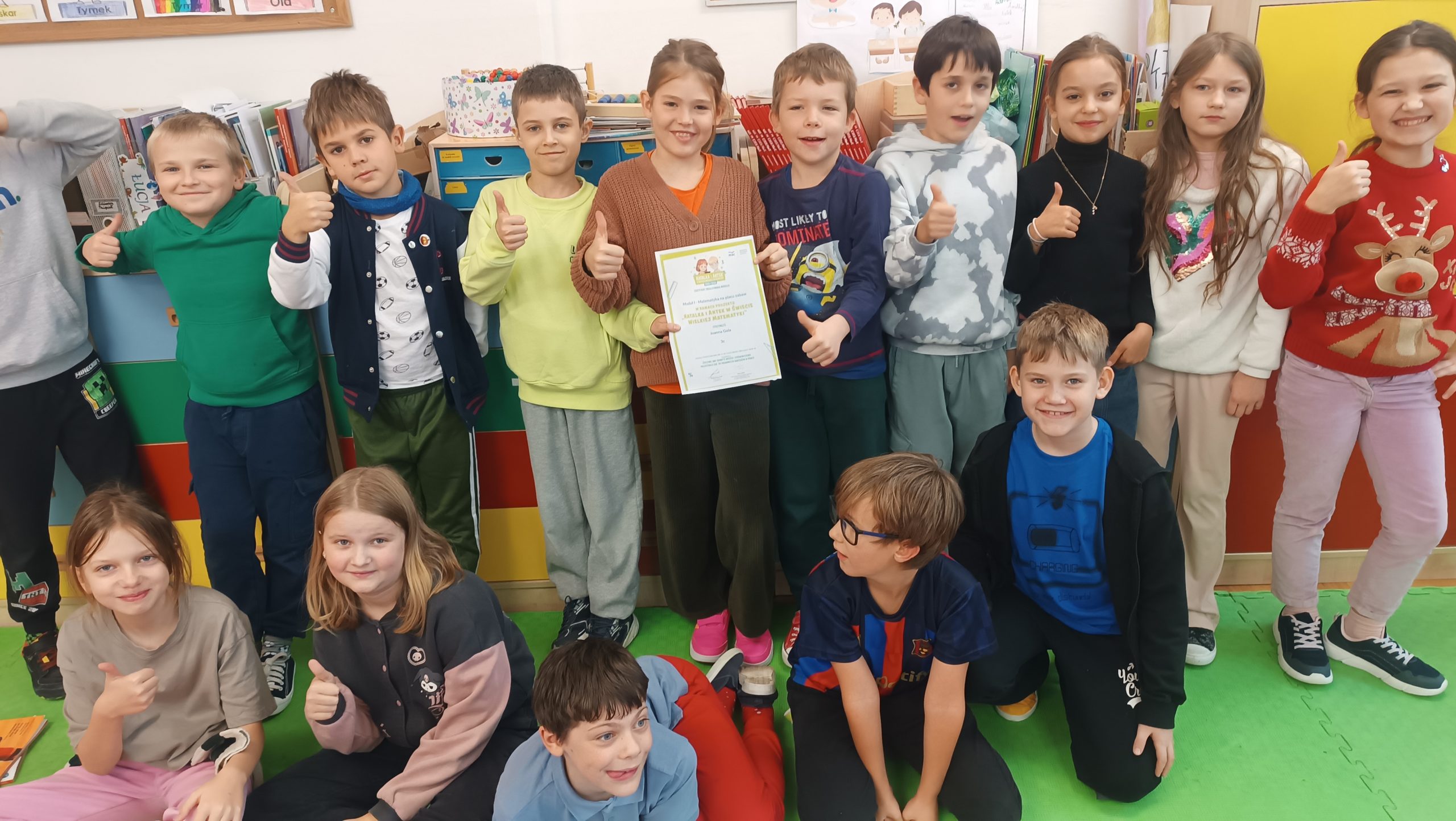 Grupa uśmiechniętych dzieci pozuje z certyfikatem ukończenia projektu,,Natalka i Antek w świecie wielkiej matematyki''. Dzieci pokazują kciuki w górę na znak radości i dumy z osiągnięć.