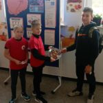 Na półpiętrze szkolnego korytarza na tle tablicy z plakatem na temat udaru mózgu stoi 2 członków Koła PP HOPR w czerwonych koszulkach. Jeden uczeń trzyma tacę z orzechami, uczennica trzyma ulotki. Przed nimi stoi inny uczeń częstujący się orzechami