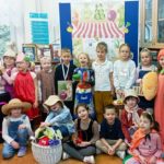 15 uczniów klas pierwszych ustawionych w dwa rzędy stoi lub siedzi na tle tablicy z regulaminem konkursu recytatorskiego oraz dekoracją przedstawiającą warzywny stragan. Dzieci trzymają w rękach owocowe i warzywne rekwizyty.