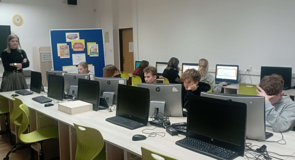 W sali komputerowej na tle plakatu reklamującego Olimpiadę Zdrowia PCK przy komputerach siedzi 8 uczniów klas starszych. Przy tablicy stoi wicedyrektorka szkoły.