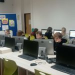 W sali komputerowej na tle plakatu reklamującego Olimpiadę Zdrowia PCK przy komputerach siedzi 8 uczniów klas starszych. Przy tablicy stoi wicedyrektorka szkoły.