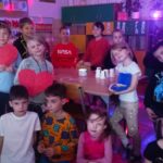 13 uczniów klasy 1c stoi w sali w koło stolika na tle kolorowej poświaty kuli disco. Na stoliku i w ich dłoniach znajdują się andrzejkowe gadżety do wróżb.