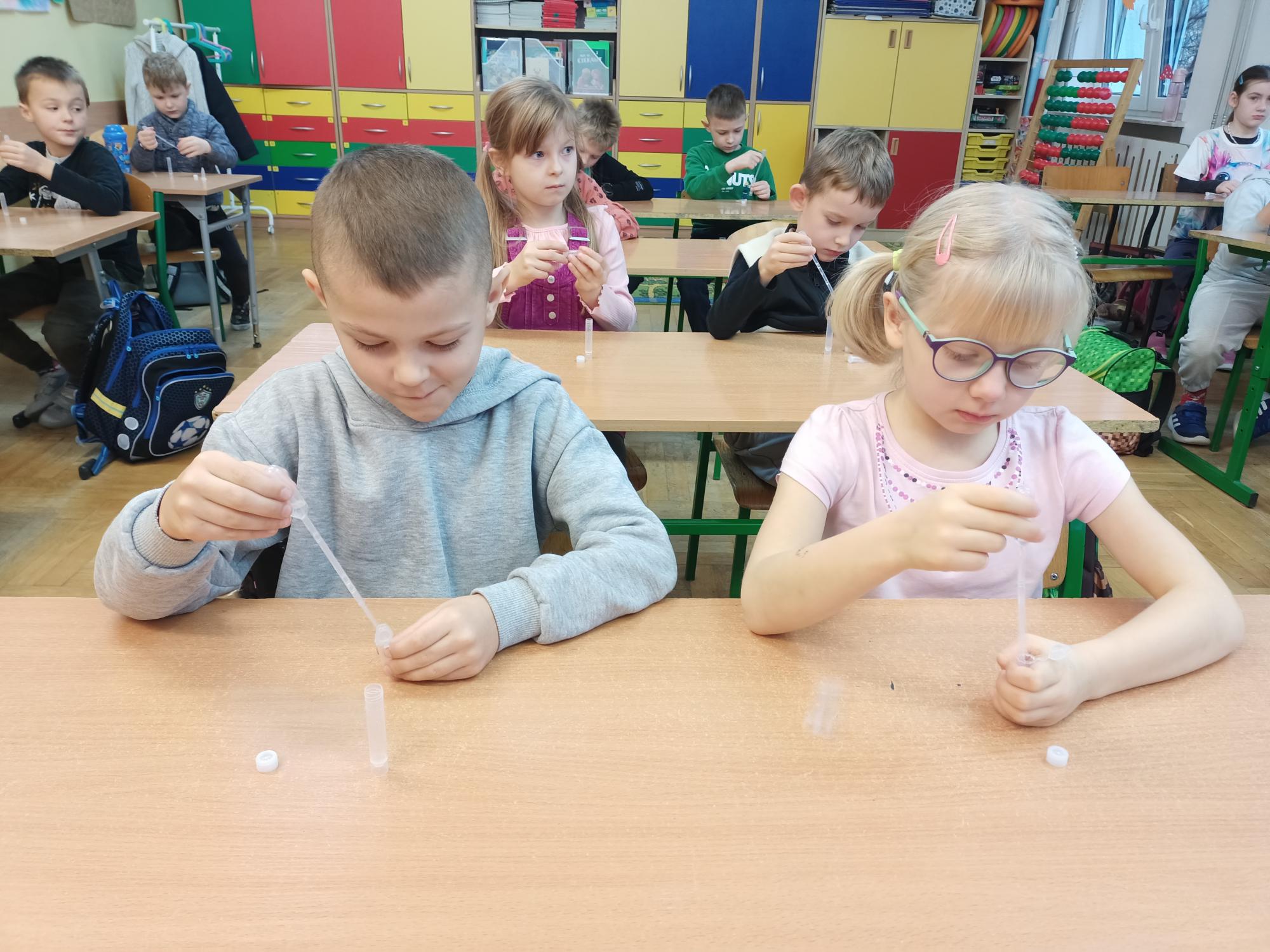 Dzieci siedzą w ławkach w sali lekcyjnej. Każde z nich przeprowadza doświadczenie. Pipetą dawkują ciecz do próbówki z nadmanganianem potasu i obserwują reakcję. 