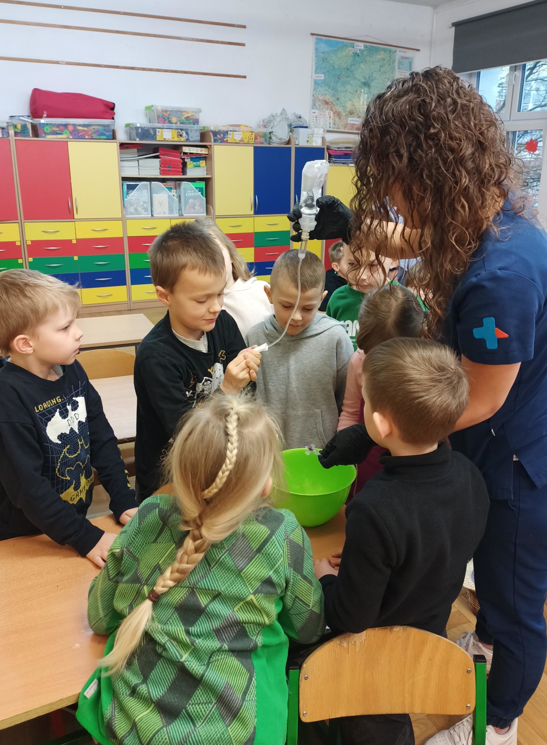 Grupa chłopców stoi obok pani weterynarz, która pokazuje im kroplówkę i tłumaczy sposób jej działania.