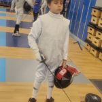 Na zdjęciu widać młodego szermierza w pełnym stroju ochronnym, stojącego na planszy tuż przed startem walki. Trzyma w dłoni maskę oraz floret, a jego postawa pokazuje skupienie i gotowość do rywalizacji. W tle widać innych zawodników przygotowujących się do swoich pojedynków