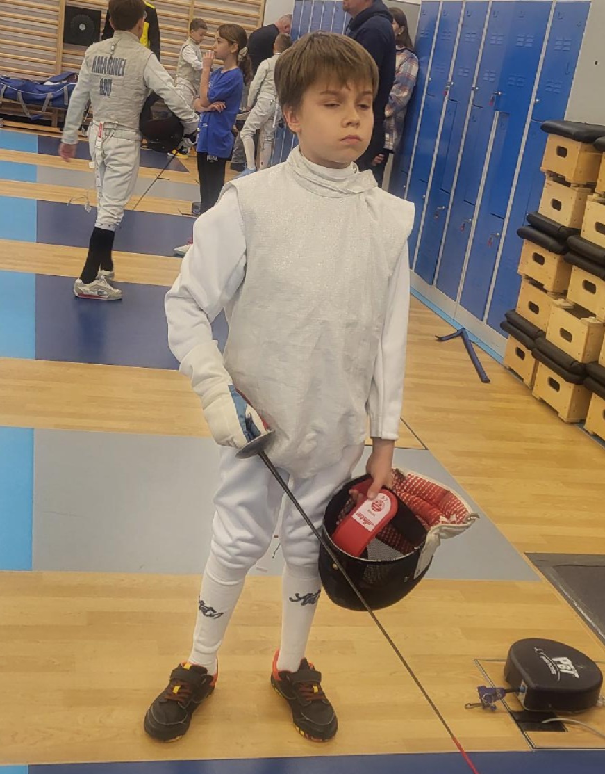 Na zdjęciu widać młodego szermierza w pełnym stroju ochronnym, stojącego na planszy tuż przed startem walki. Trzyma w dłoni maskę oraz floret, a jego postawa pokazuje skupienie i gotowość do rywalizacji. W tle widać innych zawodników przygotowujących się do swoich pojedynków