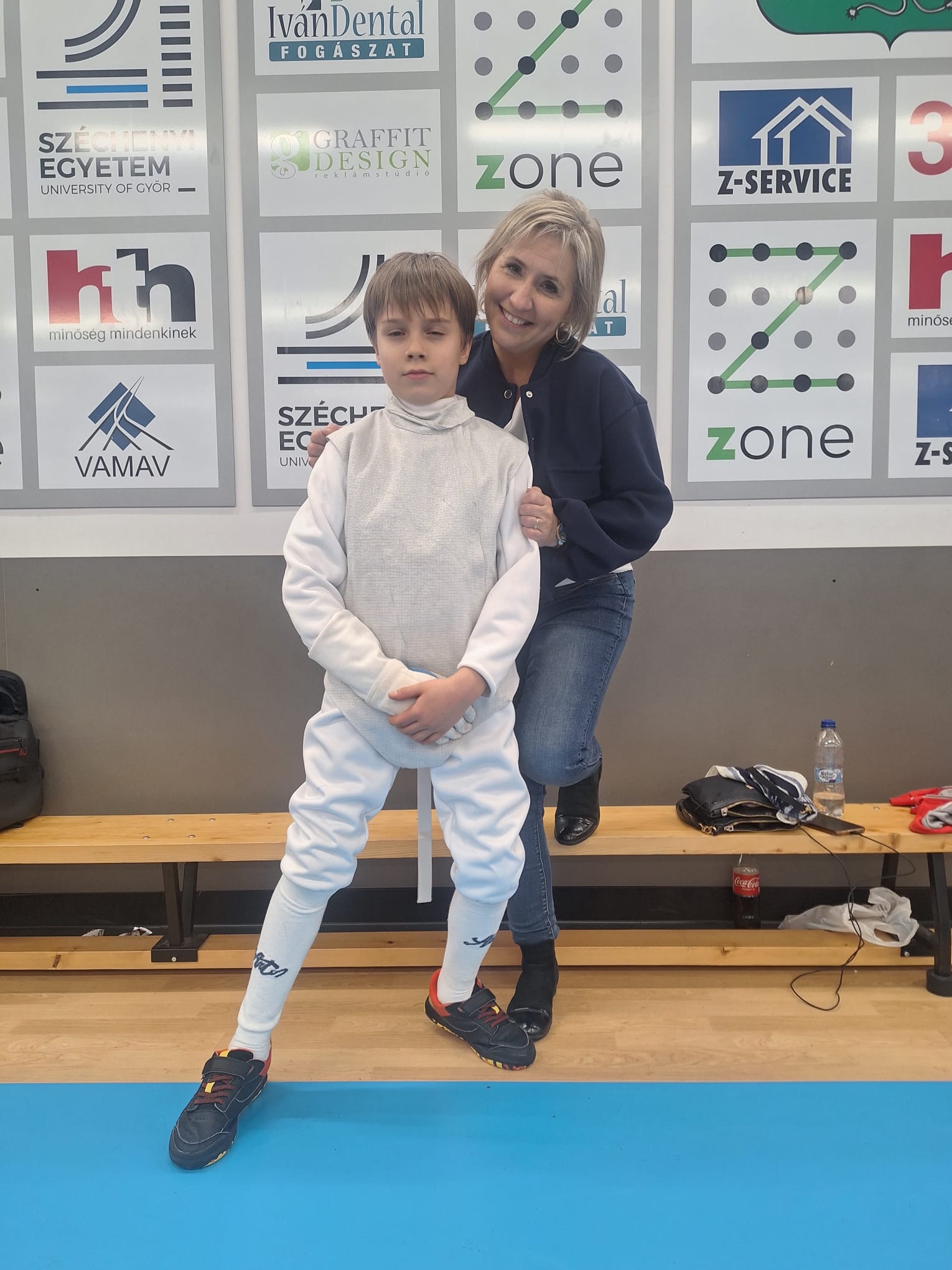 Na zdjęciu znajduje się uczeń klasy 3 w stroju szermierczym, stojący obok swojej nauczycielki. Oboje pozują z uśmiechem na tle tablicy z logotypami, co tworzy miłą i wspierającą atmosferę. Uczeń wygląda na dumnego ze swojego udziału w zawodach, a nauczycielka towarzyszy mu z widoczną życzliwością i wsparciem.