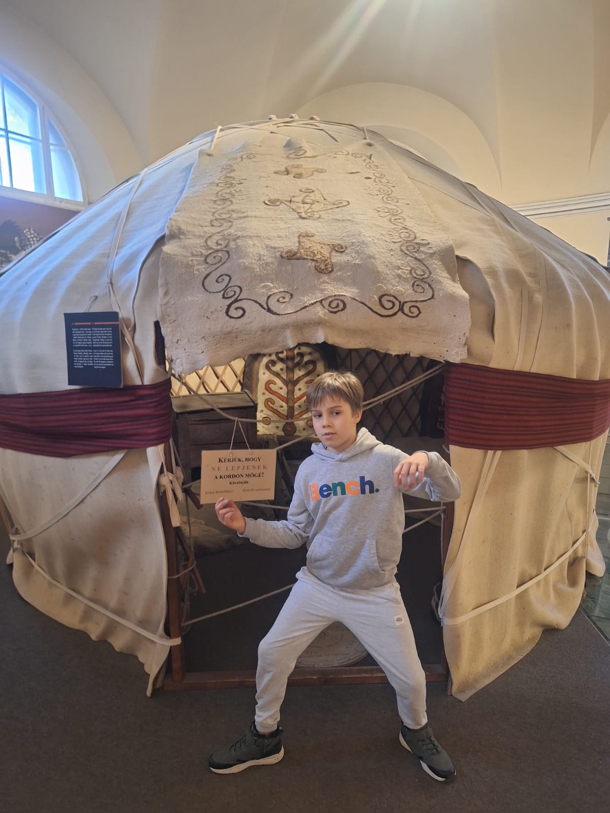 Na zdjęciu widoczny jest uczeń klasy 3, który pozuje przed tradycyjną mongolską jurtą znajdującą się w muzeum. Ubrany w sportowy dres, przyjmuje dynamiczną pozę, jakby nawiązywał do swoich szermierczych umiejętności. Tło stanowi efektowna konstrukcja jurty wraz z jej dekoracyjnymi detalami, co nadaje zdjęciu ciekawy, edukacyjny charakter.