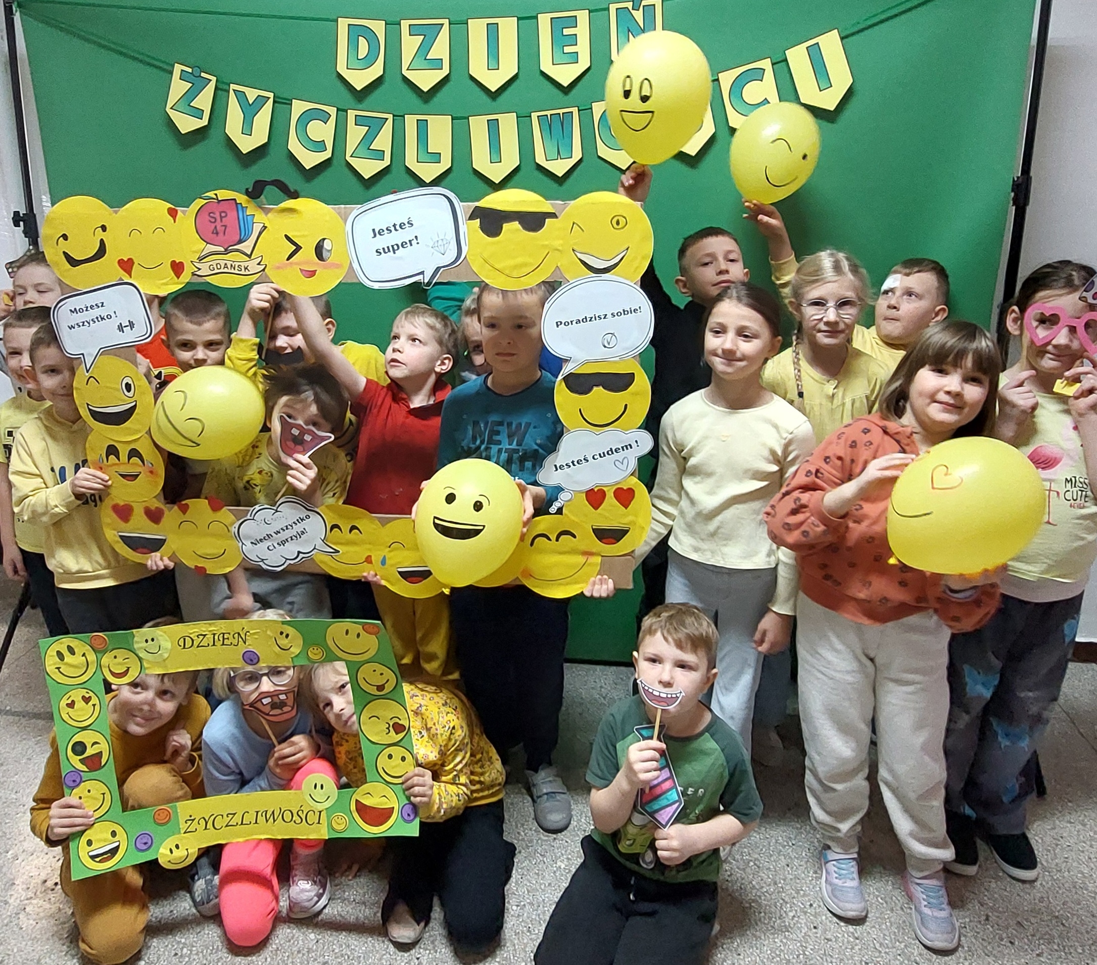 Grupa dzieci ubranych na żółto pozuje z papierowymi emotikonami, balonami i akcesoriami do zdjęć z okazji „Dnia Życzliwości”, stojąc przed zielonym tłem z dekoracyjnym napisem DZIEŃ ŻYCZLIWOŚCI