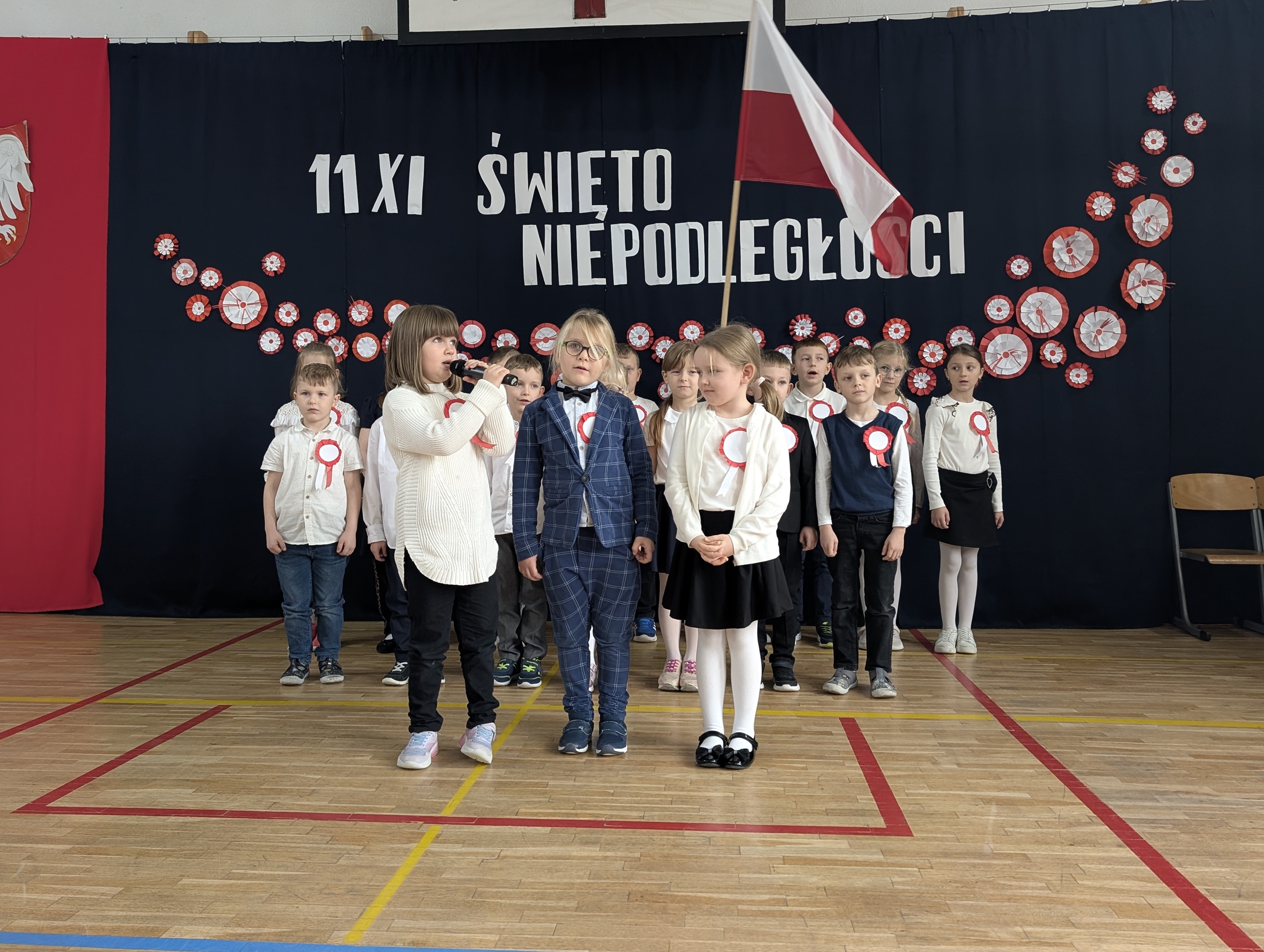 Grupa uczniów z klasy 1a stoi na sali gimnastycznej przed dekoracją z napisem „11 XI Święto Niepodległości”. Dzieci ubrane są odświętnie, mają przyczepione do ubrań kokardy narodowe.