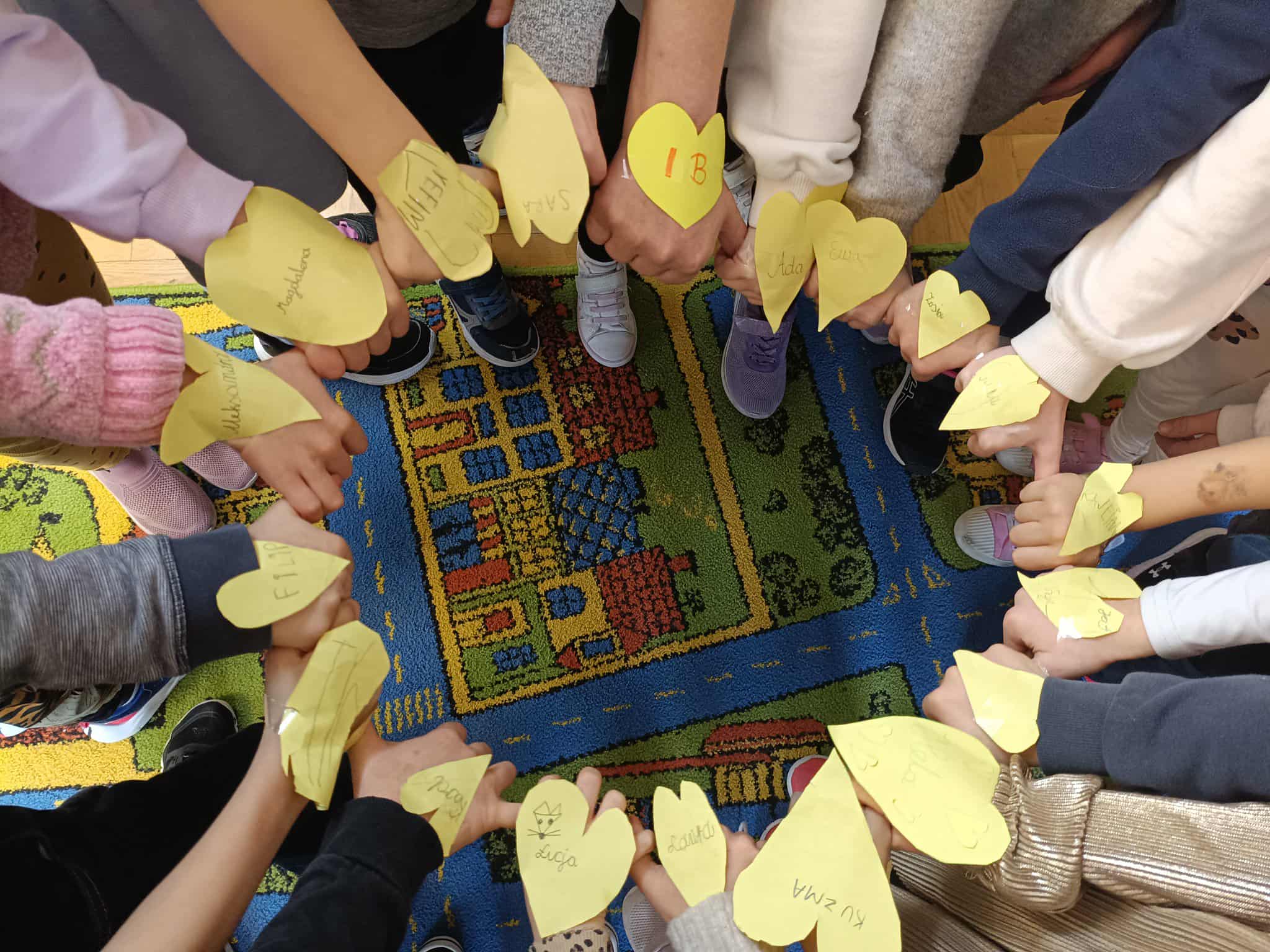 Dzieci stoją w kręgu i trzymają nad dywanem żółte papierowe serduszka z imionami oraz miłymi hasłami.