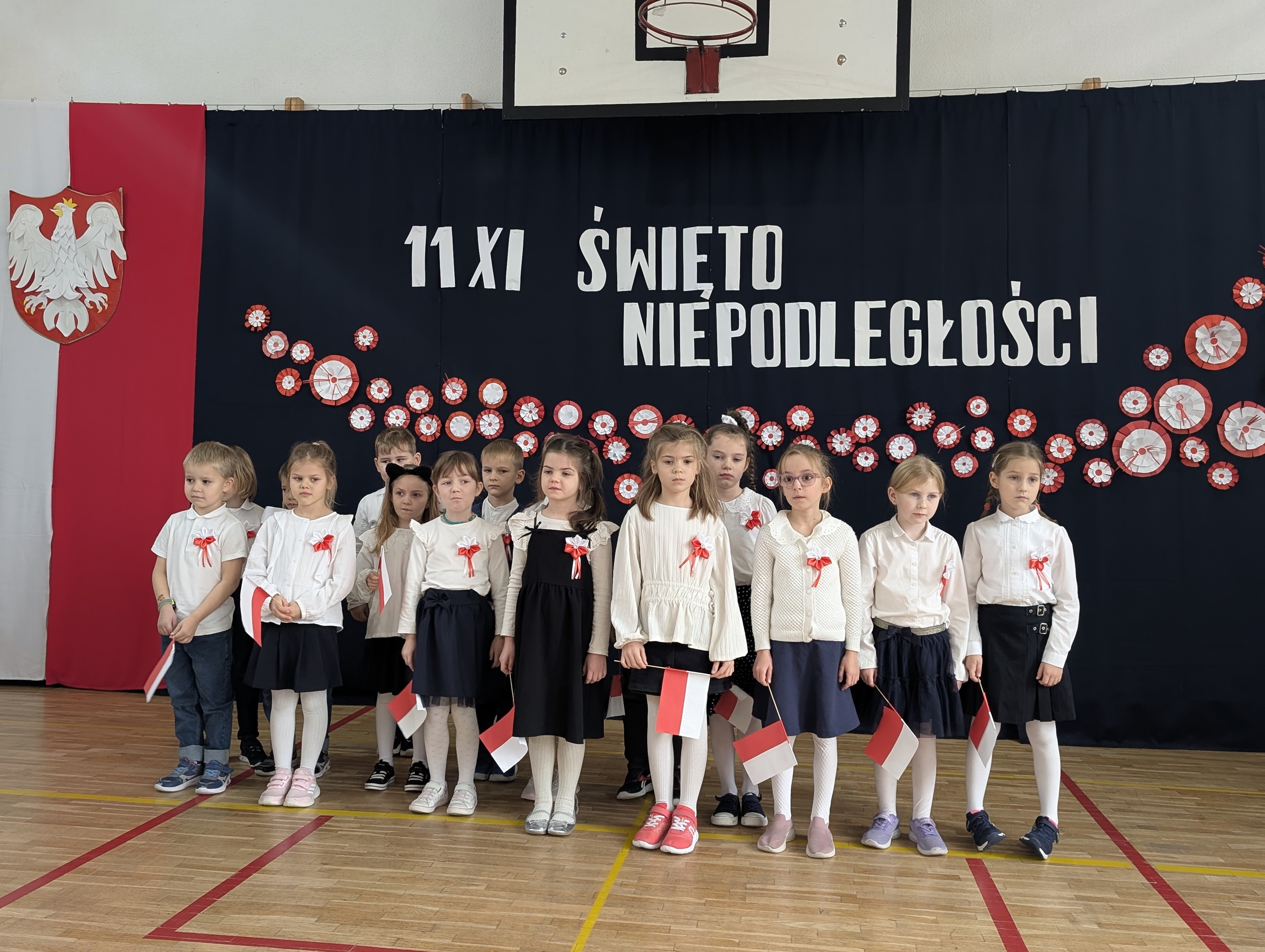 Grupa uczniów z klasy 1b stoi na sali gimnastycznej przed dekoracją z napisem „11 XI Święto Niepodległości”. Dzieci ubrane są odświętnie, mają przyczepione do ubrań kokardy narodowe, a w rękach trzymają flagi.