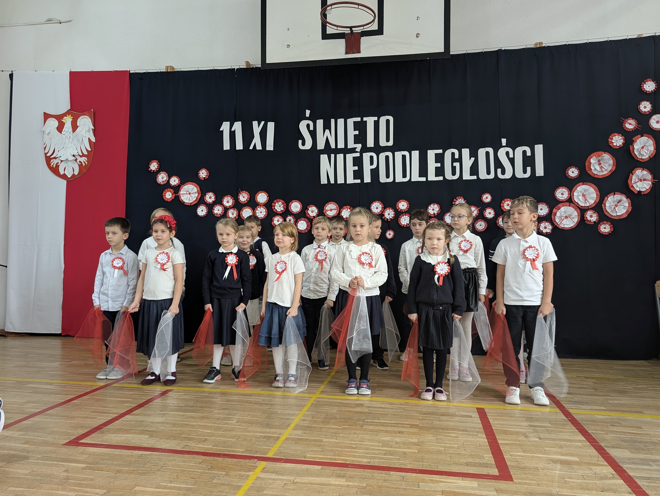 Uczniowie klasy 1c stoją na tle dekoracji z okazji Święta Niepodległości. Mają biało-czerwone kokardy i trzymają w rękach czerwone oraz białe wstążki z tiulu.