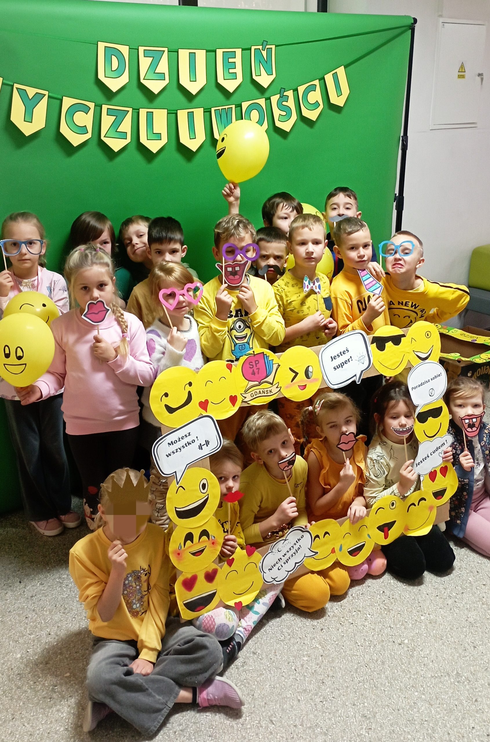 Grupa dzieci ubranych na żółto pozuje z papierowymi emotikonami, balonami i akcesoriami do zdjęć z okazji „Dnia Życzliwości”, stojąc przed zielonym tłem z dekoracyjnym napisem DZIEŃ ŻYCZLIWOŚCI