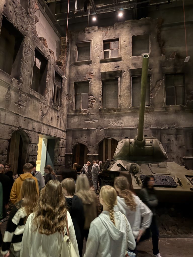 Grupa uczniów zwiedza ekspozycję przedstawiającą zniszczoną, powojenną ulicę. W tle widać wysokie, zrujnowane kamienice z wybitymi oknami i odsłoniętą cegłą. Po prawej stronie stoi duży czołg.