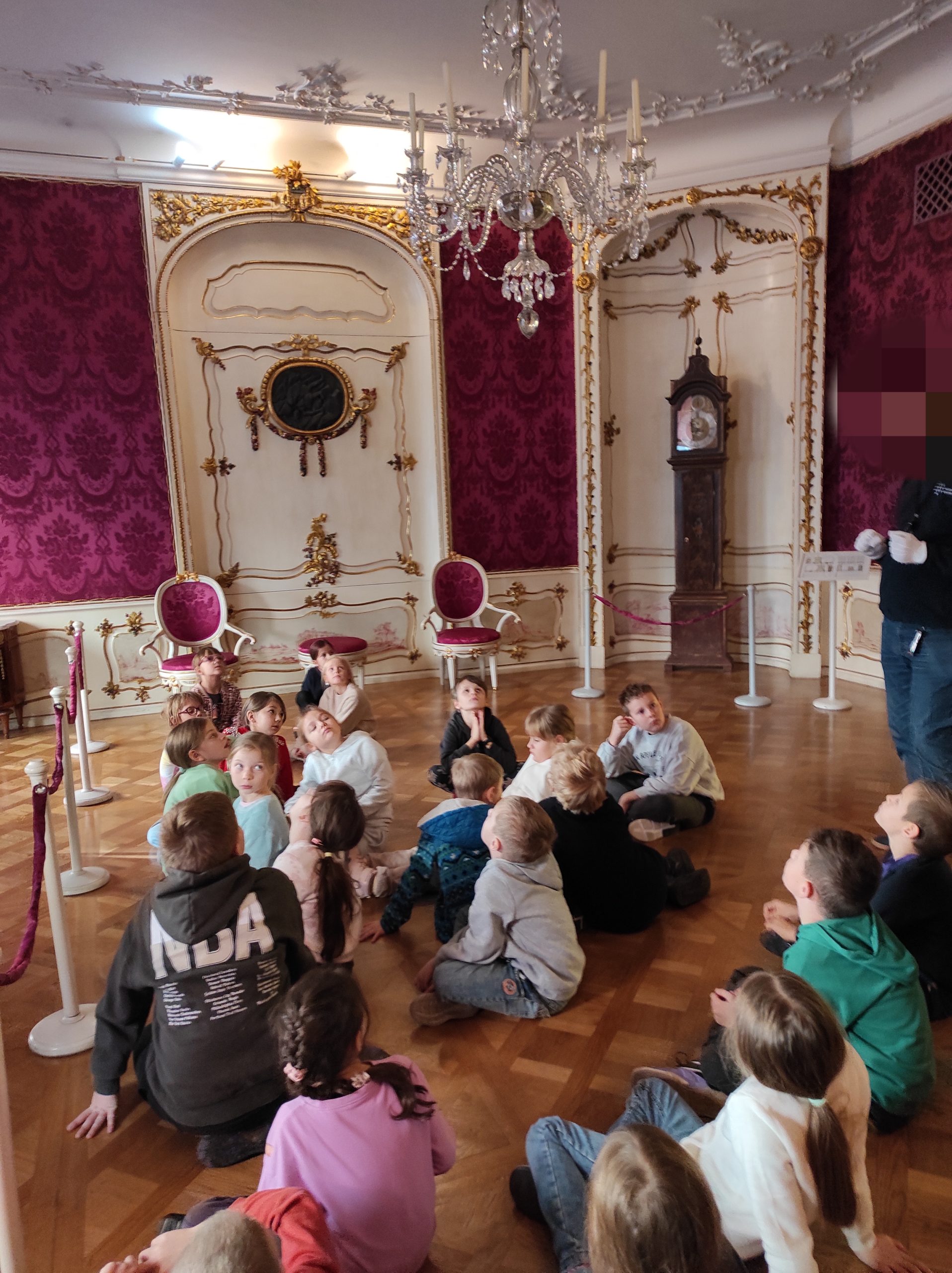 22 uczniowie siedzą na podłodze w strojnie urządzonej sali. Przy suficie widoczny kryształowy żyrandol, na podłodze parkiet, na ścianach satynowe tapety.