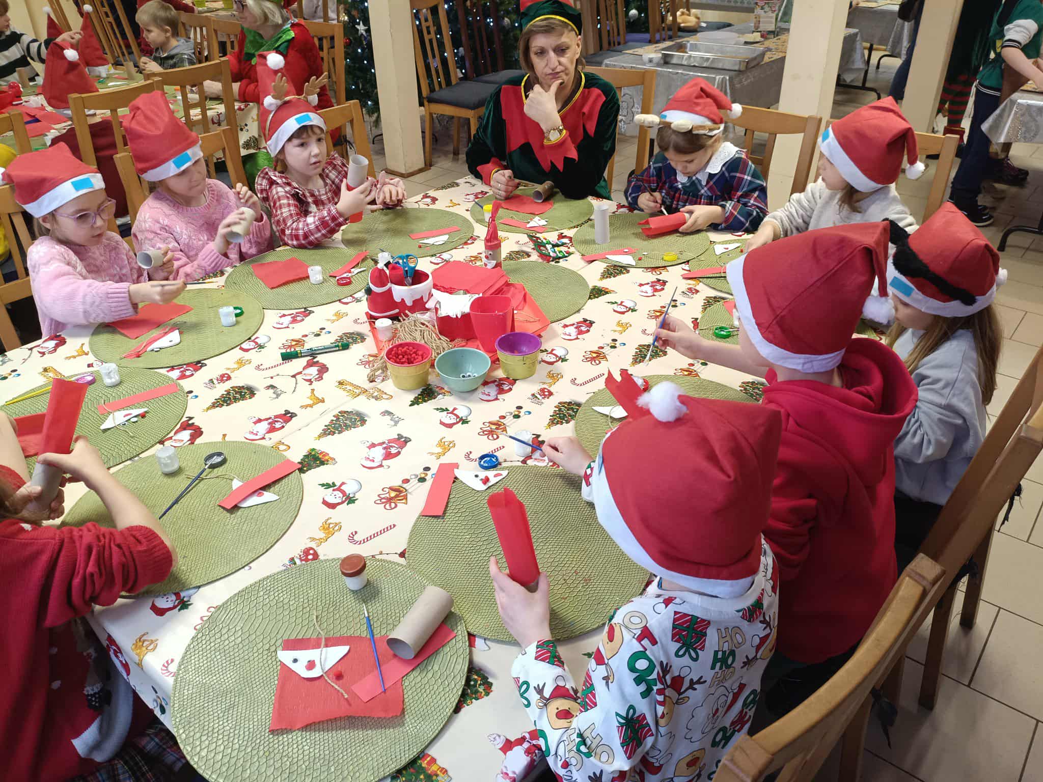 Dzieci w czapkach Mikołaja siedzą wokół stołu, wykonując świąteczne ozdoby z papieru i rolek kartonowych. Na stole leżą kolorowe przybory, miseczki z materiałami i zielone podkładki. Przy dzieciach stoi osoba przebrana za elfa, nadzorująca zajęcia.