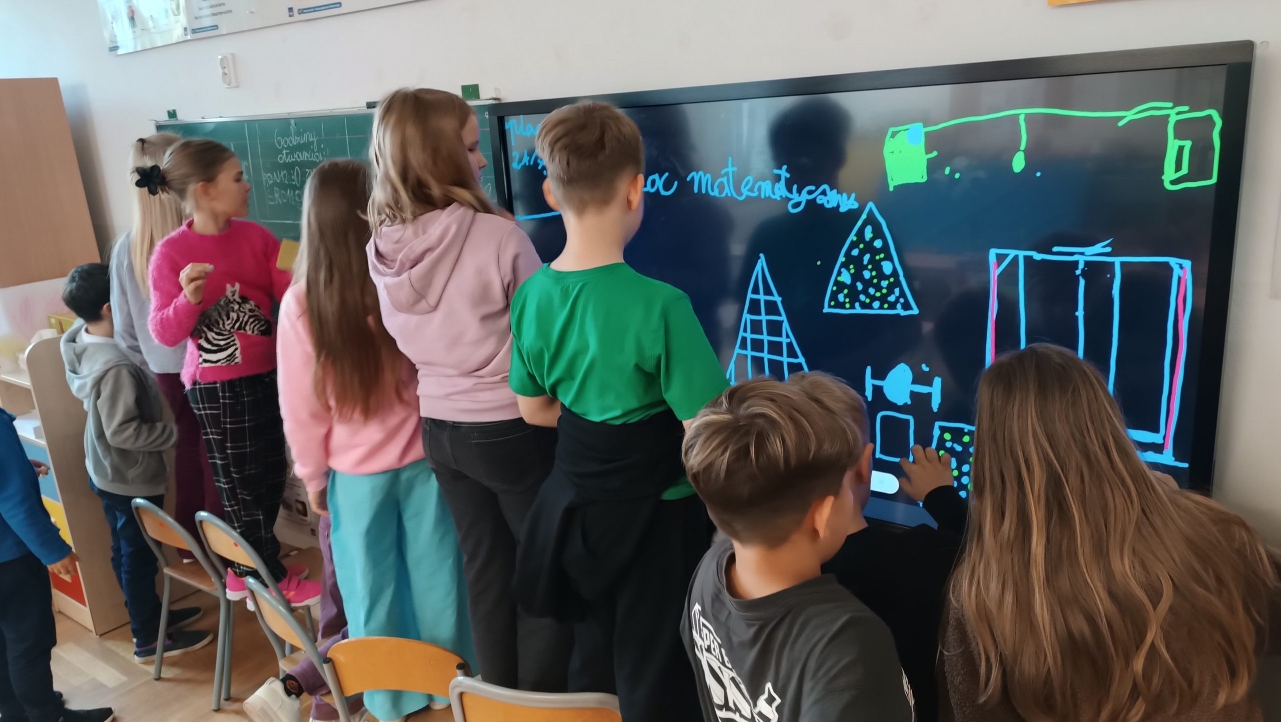  Grupa dzieci pracuje przy tablicy interaktywnej, rysuąc i omawiając projekt,,Placu matematycznego''.Uczniowie stoją przy tablicy, na której widnieją kolorowe kształty i napisy.