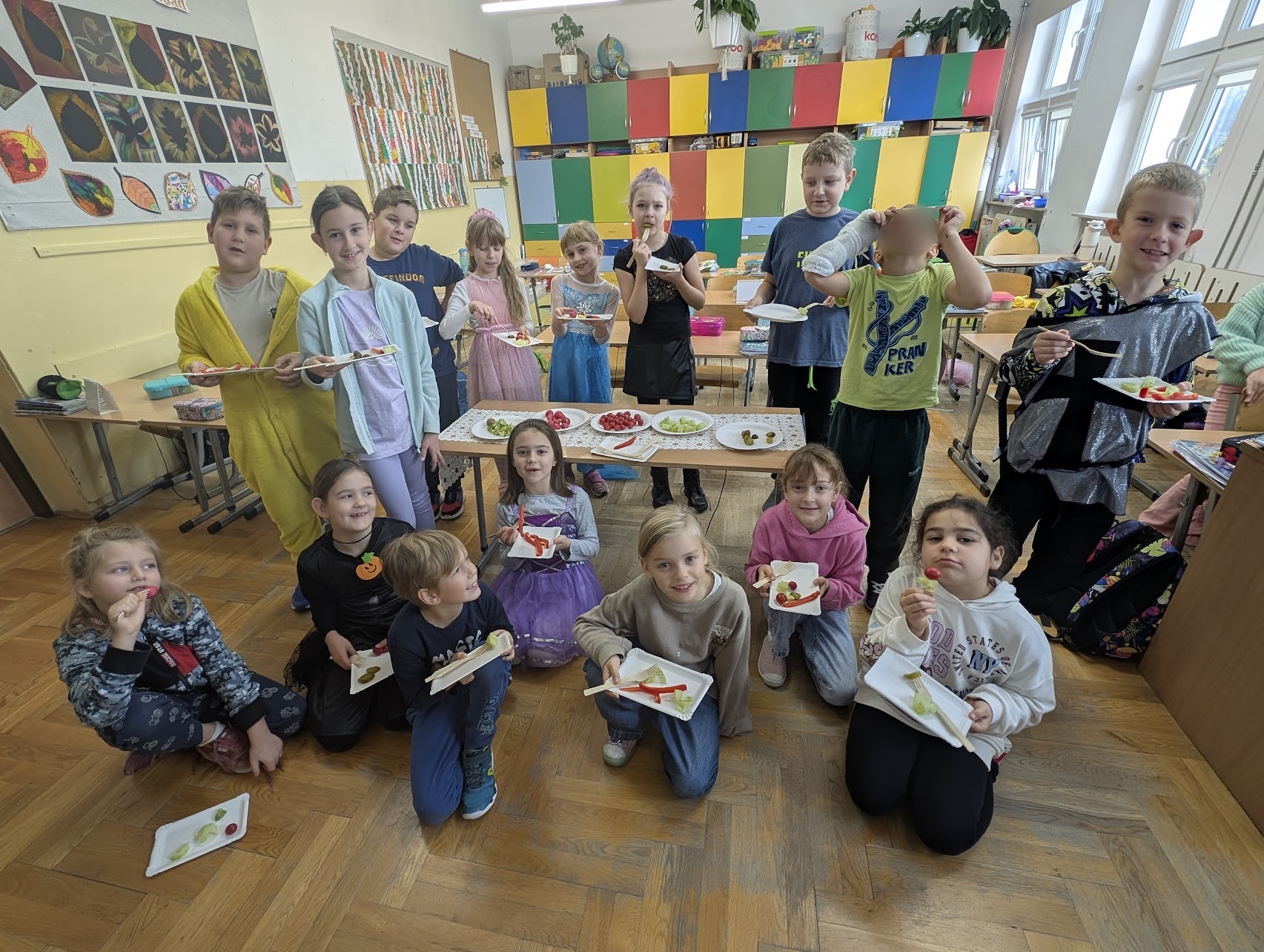 Grupa dzieci z 2c pozuje w klasie szkolnej, stojąc i siedząc w kilku rzędach. Trzymają papierowe talerzyki z warzywami, wyglądając na zadowolone. W tle widać kolorowe szafki, prace plastyczne na ścianie i duże okna, przez które wpada światło dzienne.