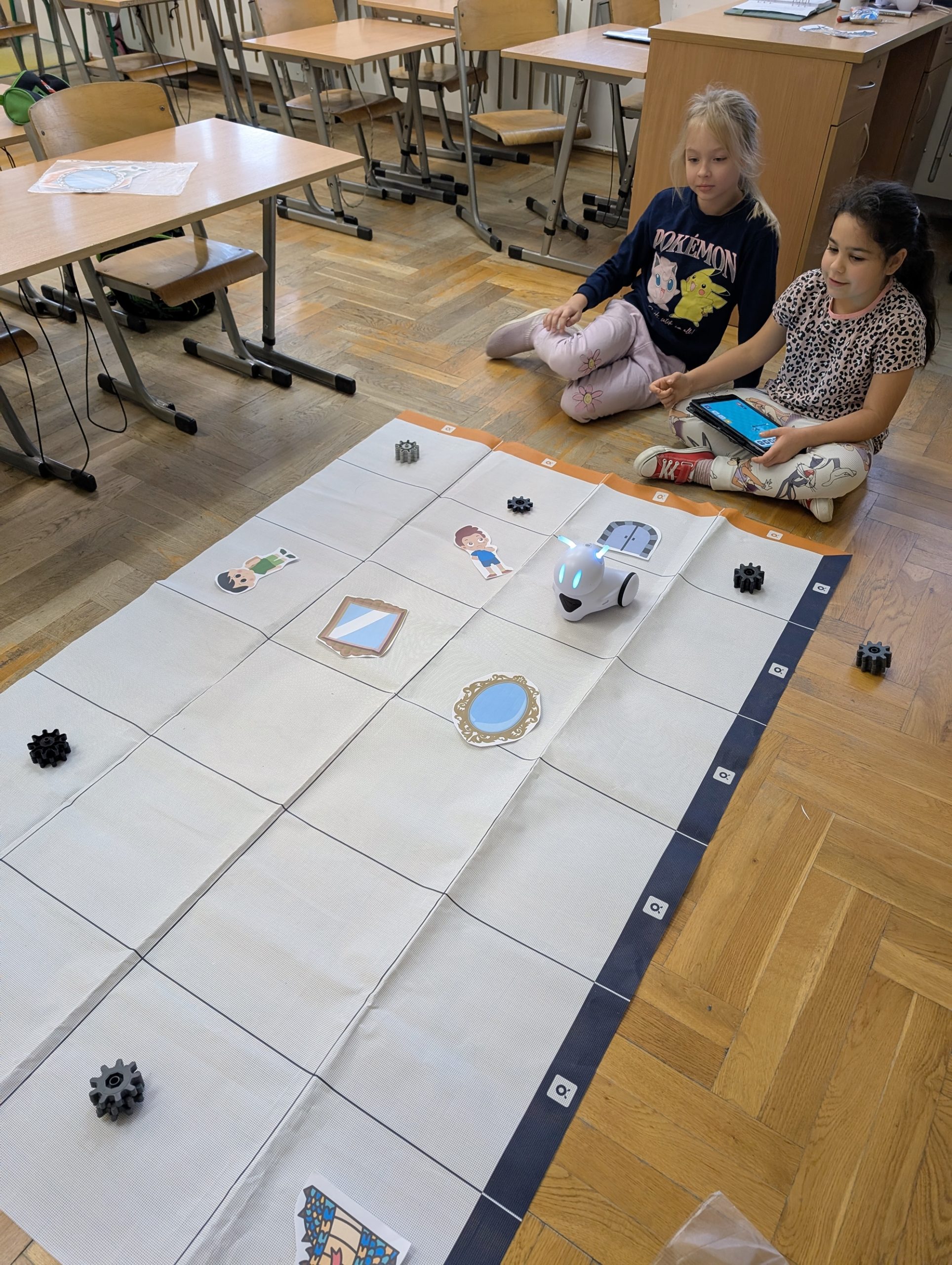 Dwie dziewczynki siedzą na podłodze w sali lekcyjnej obok dużej maty edukacyjnej z ilustracjami postaci i przedmiotów. Przed nimi znajduje się mały robot, a jedna z dziewczynek trzyma tablet.