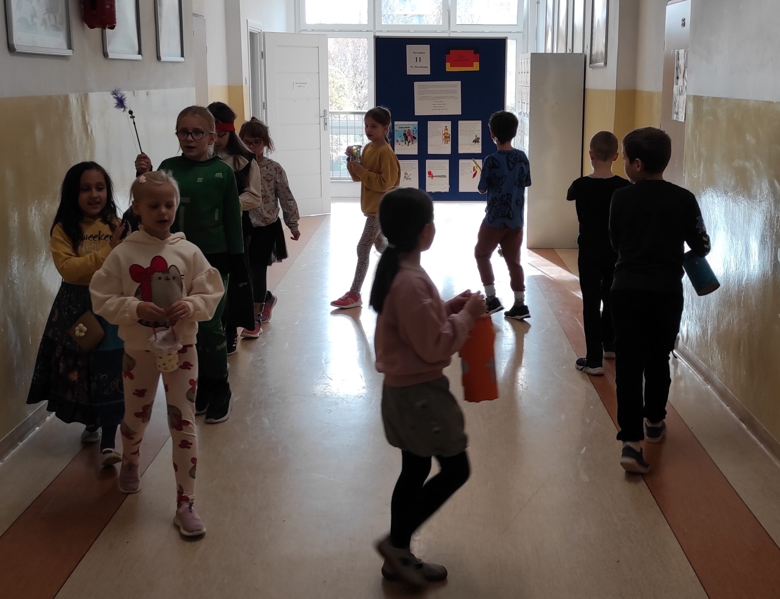 Uczniowie stoją w kręgu, na szkolnym korytarzu. W dłoniach trzymają lampiony. W tle elementy szkolnego korytarza.