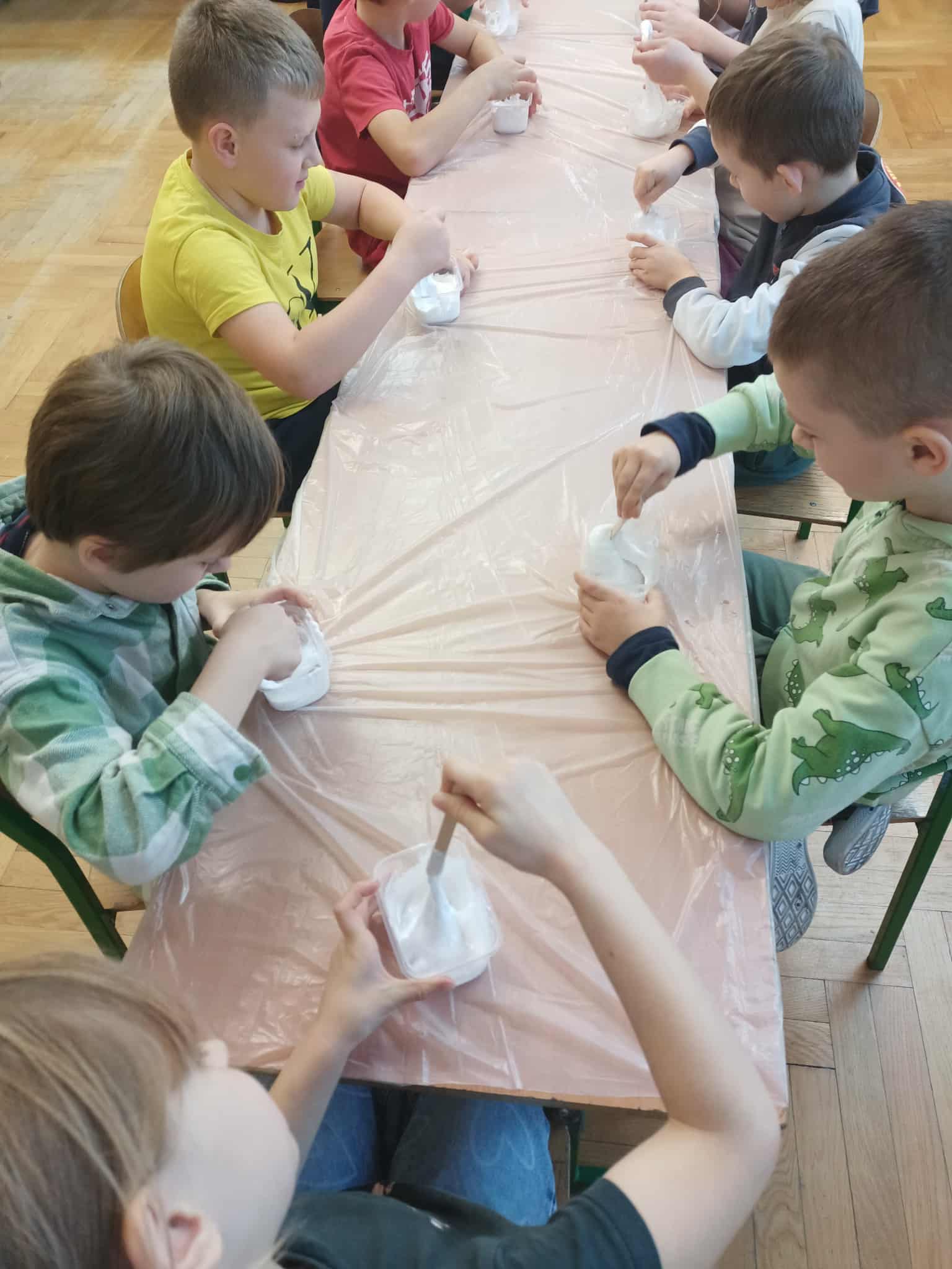 Dzieci siedzą przy długim stole zabezpieczonym folią i mieszają białą masę w plastikowych pojemnikach, przygotowując slime.