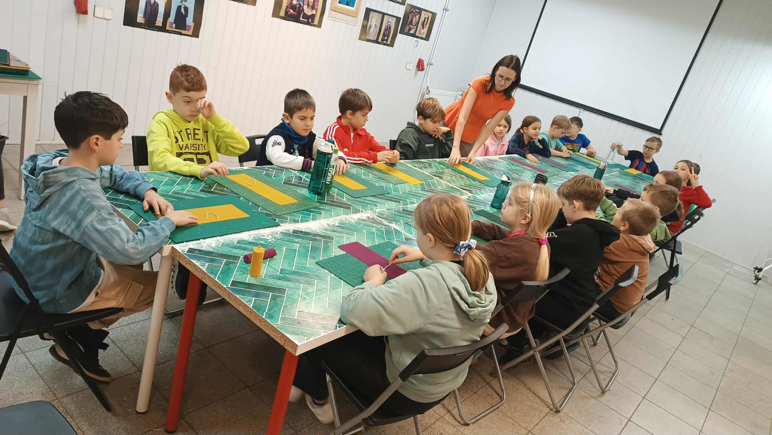 Grupa dzieci siedzi przy dużym stole wyłożonym zielonymi matami do prac plastycznych. Każde dziecko ma przed sobą arkusz węzy pszczelej. Obecna jest również osoba prowadząca warsztaty. Dzieci pracują w skupieniu, przygotowując własne świeczki.