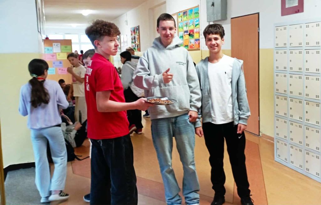 Na szkolnym korytarzu uczeń koła PP HOPR z tacą z orzechami, obok dwaj uczniowie częstujący się orzechami. W tle szafki szkolne i inni uczniowie