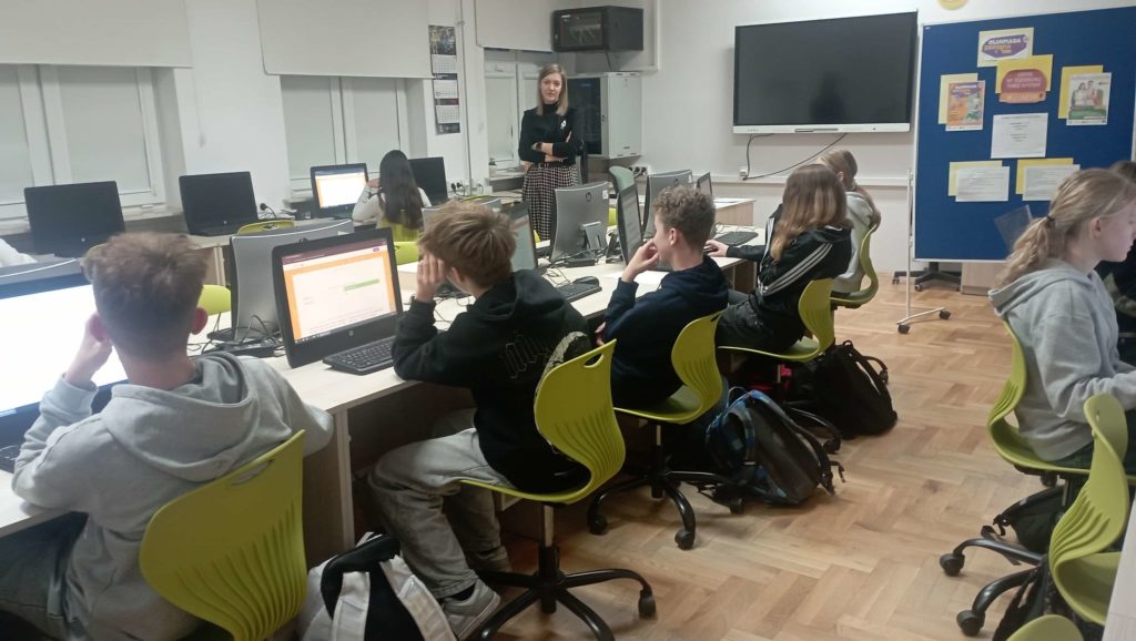 W sali komputerowej na tle tablicy multimedialnej, plakatu reklamującego Olimpiadę Zdrowia PCK oraz okien przy komputerach siedzi 7 uczniów klas starszych. W tle wicedyrektorka szkoły.