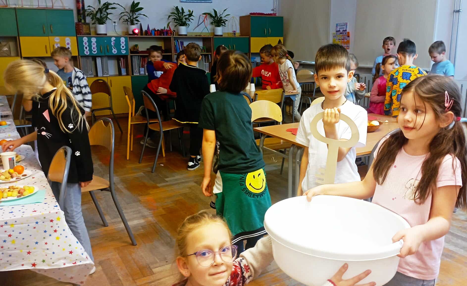 Na pierwszym planie 2 uczennice klasy 1c trzymające miskę z wodą, jeden uczeń z tekturowym kluczem oraz inny uczeń rzucający zza pleców monetą do miski. W tle inne dzieci z klasy 1c przy złączonych stolikach bawią się we wróżby oraz zajadają poczęstunek.