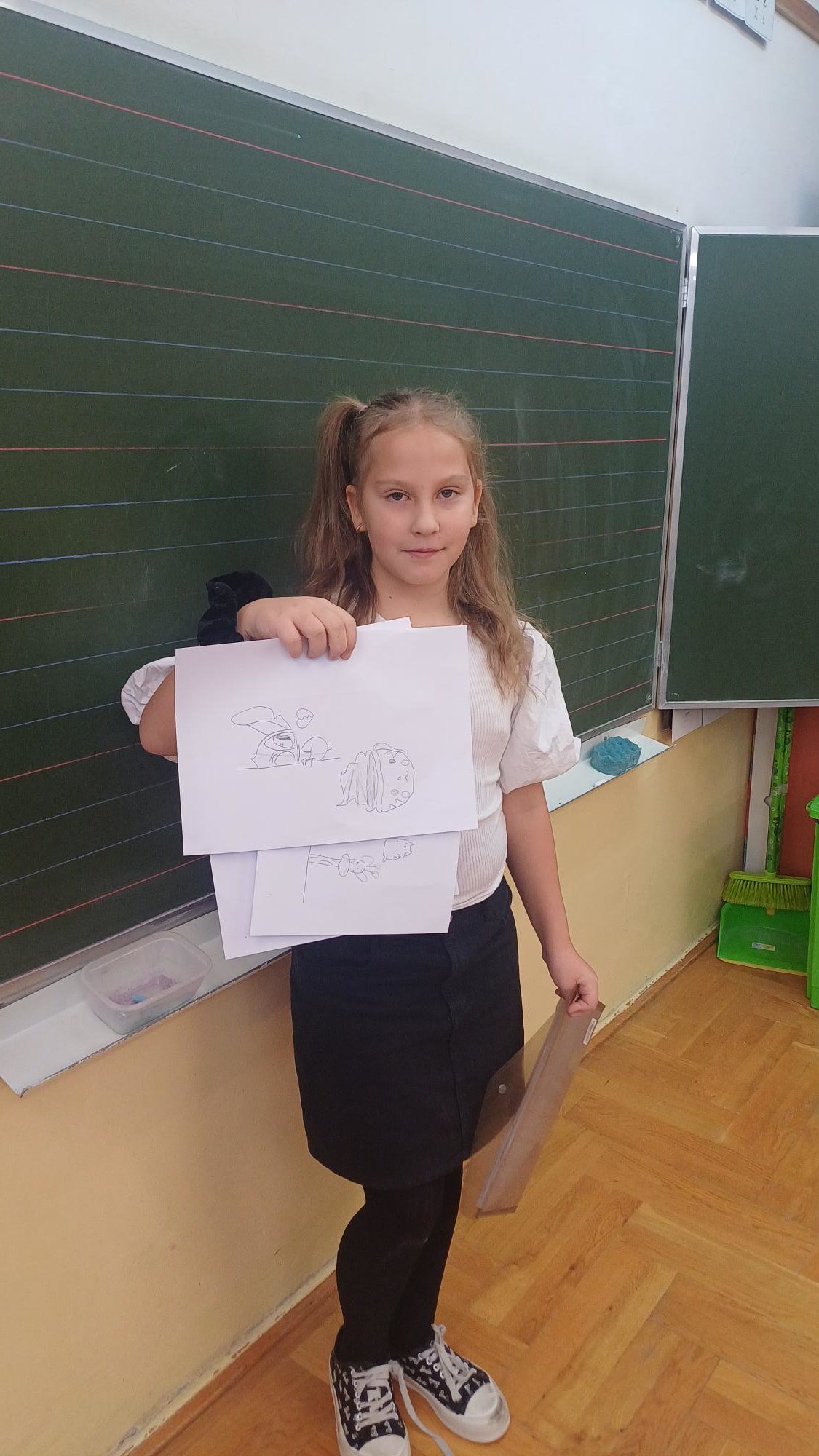 Dziewczynka stoi przed tablicą pokazując swoje rysunki 