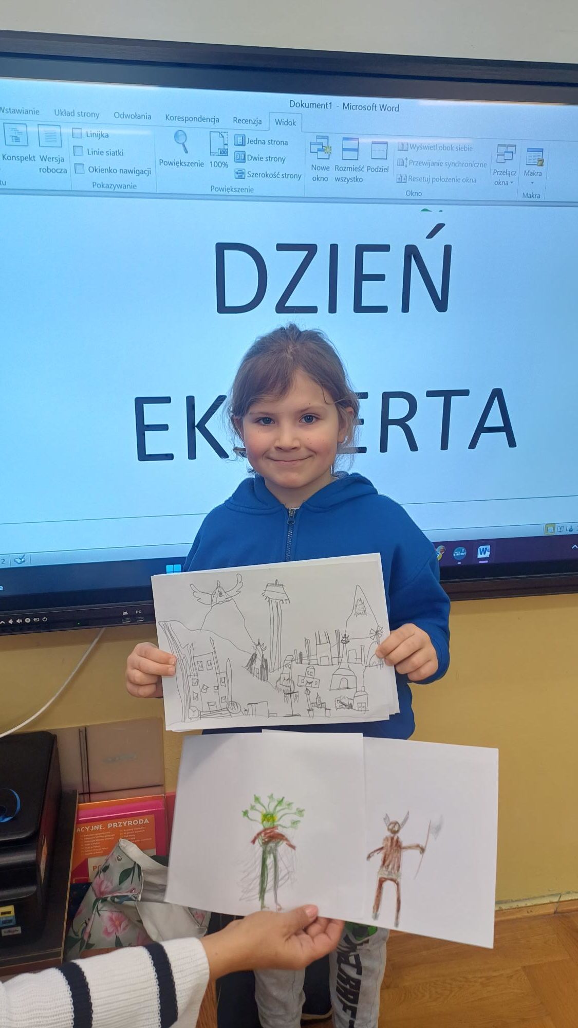 chłopiec trzyma kartkę przed tablicą, za nim napis Dzień Eksperta