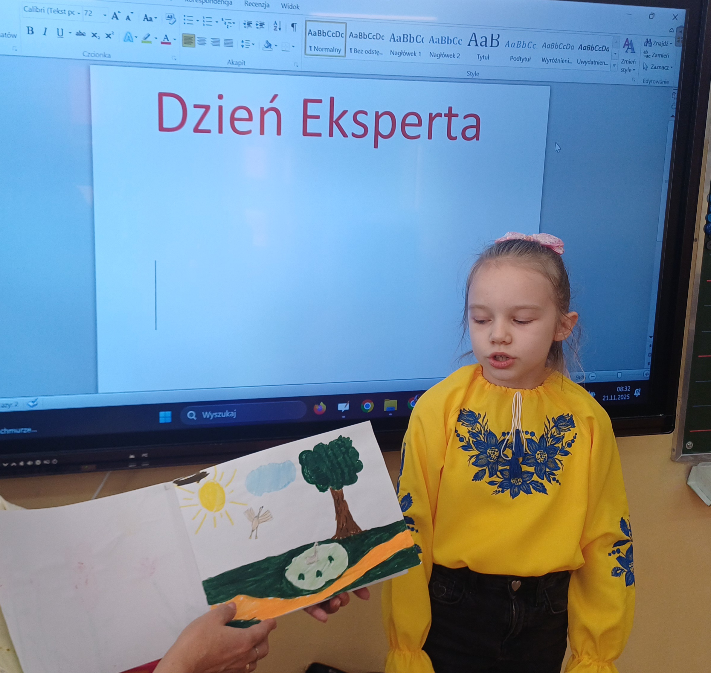 Dziewczynka w żółtej bluzce stoi przed tablicą spoglądając na blok z pracami plastycznym