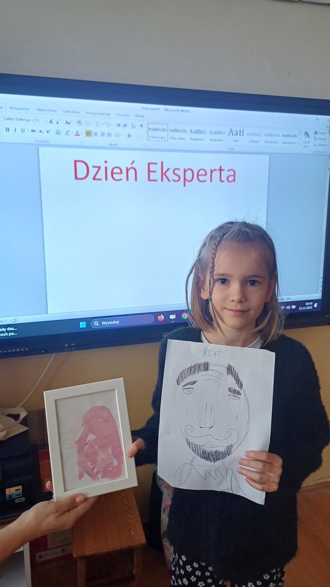 Dziewczynka w żółtej bluzce stoi przed tablicą spoglądając na blok z pracami plastycznym