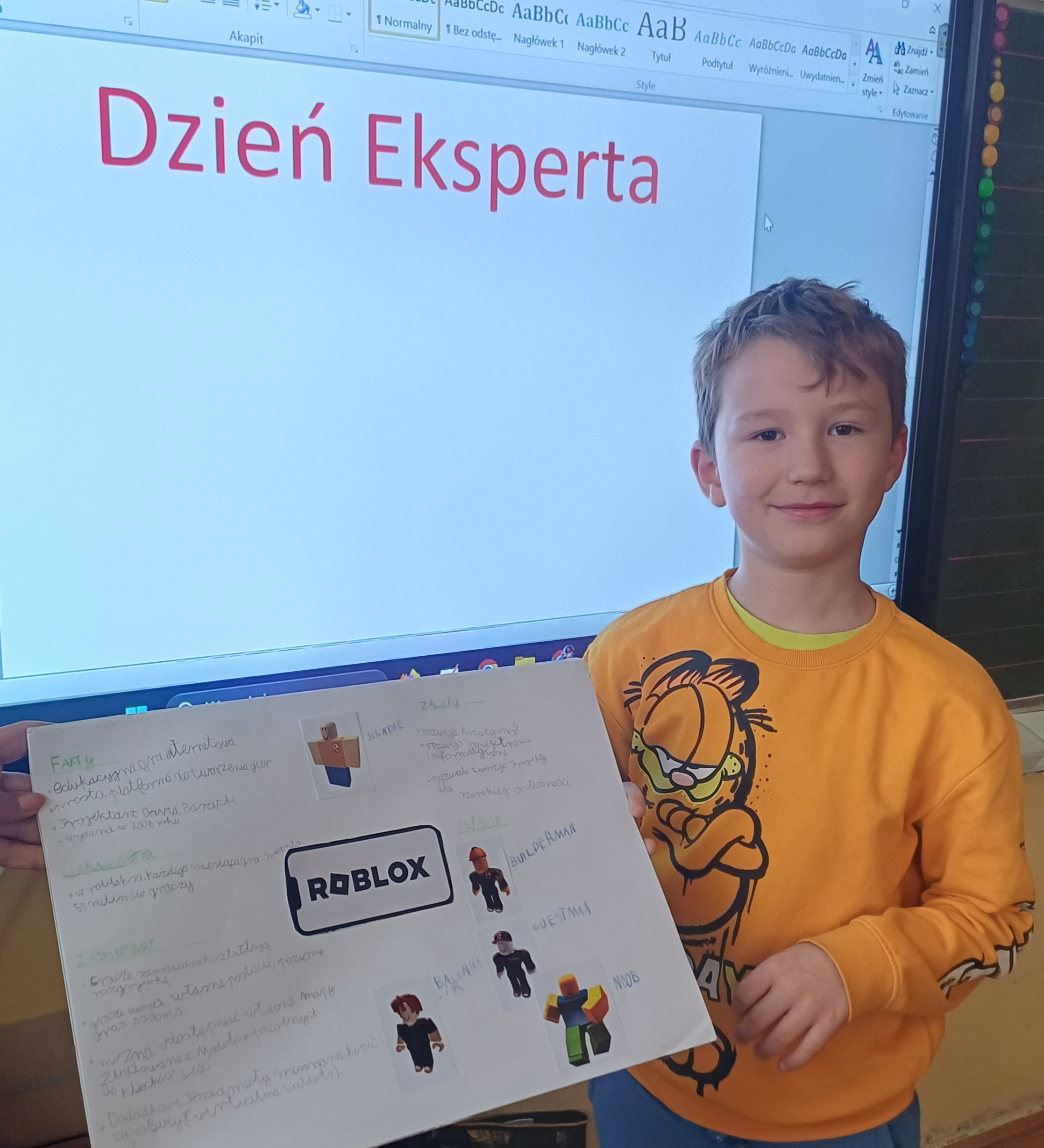 Chłopiec stoi przed tablicą, trzymając w rękach blok z rysunkami.