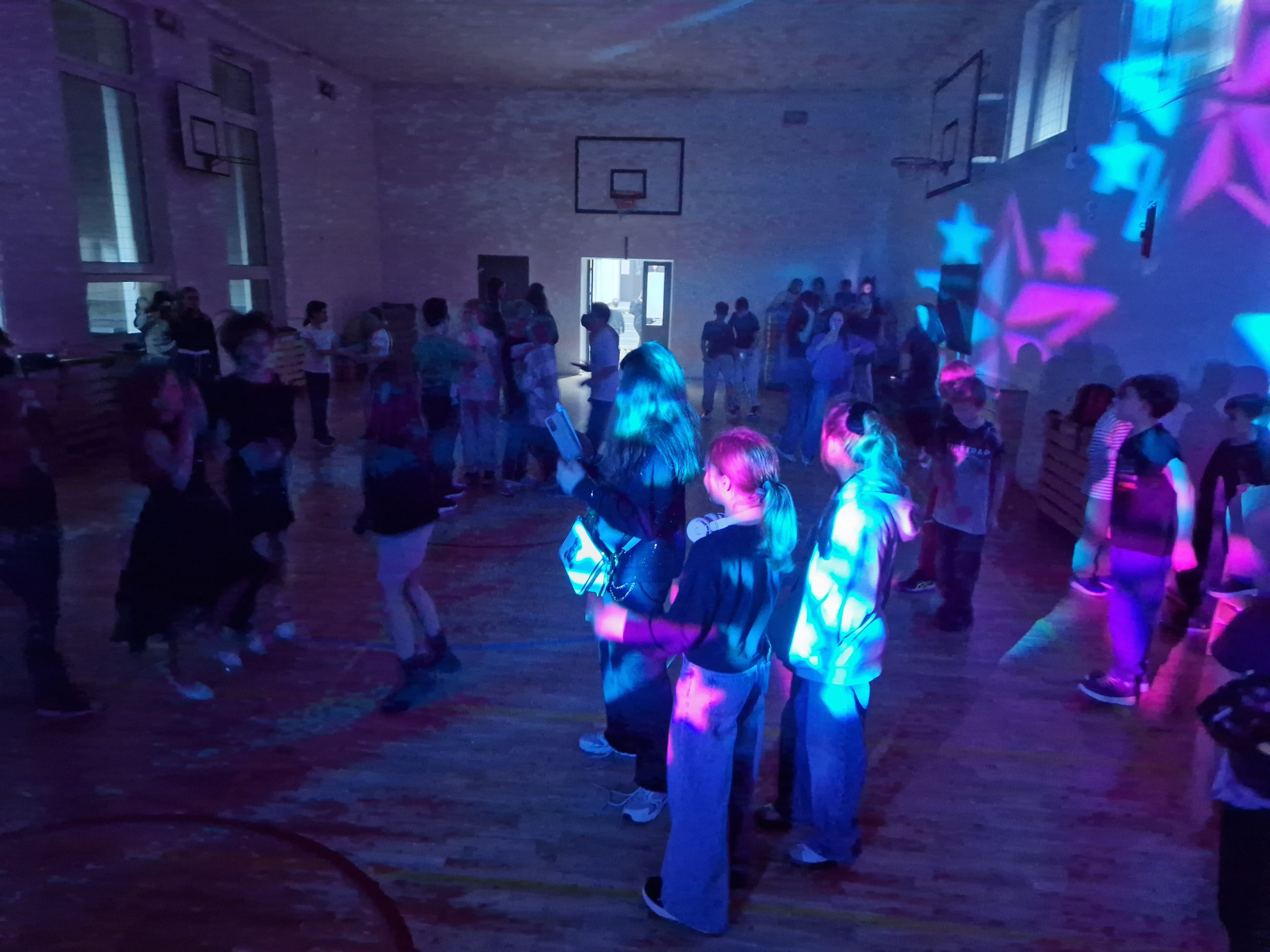 Cztery uczennice stojące razem na sali gimnastycznej podczas dyskoteki andrzejkowej, pozujące do zdjęcia w kolorowym świetle.