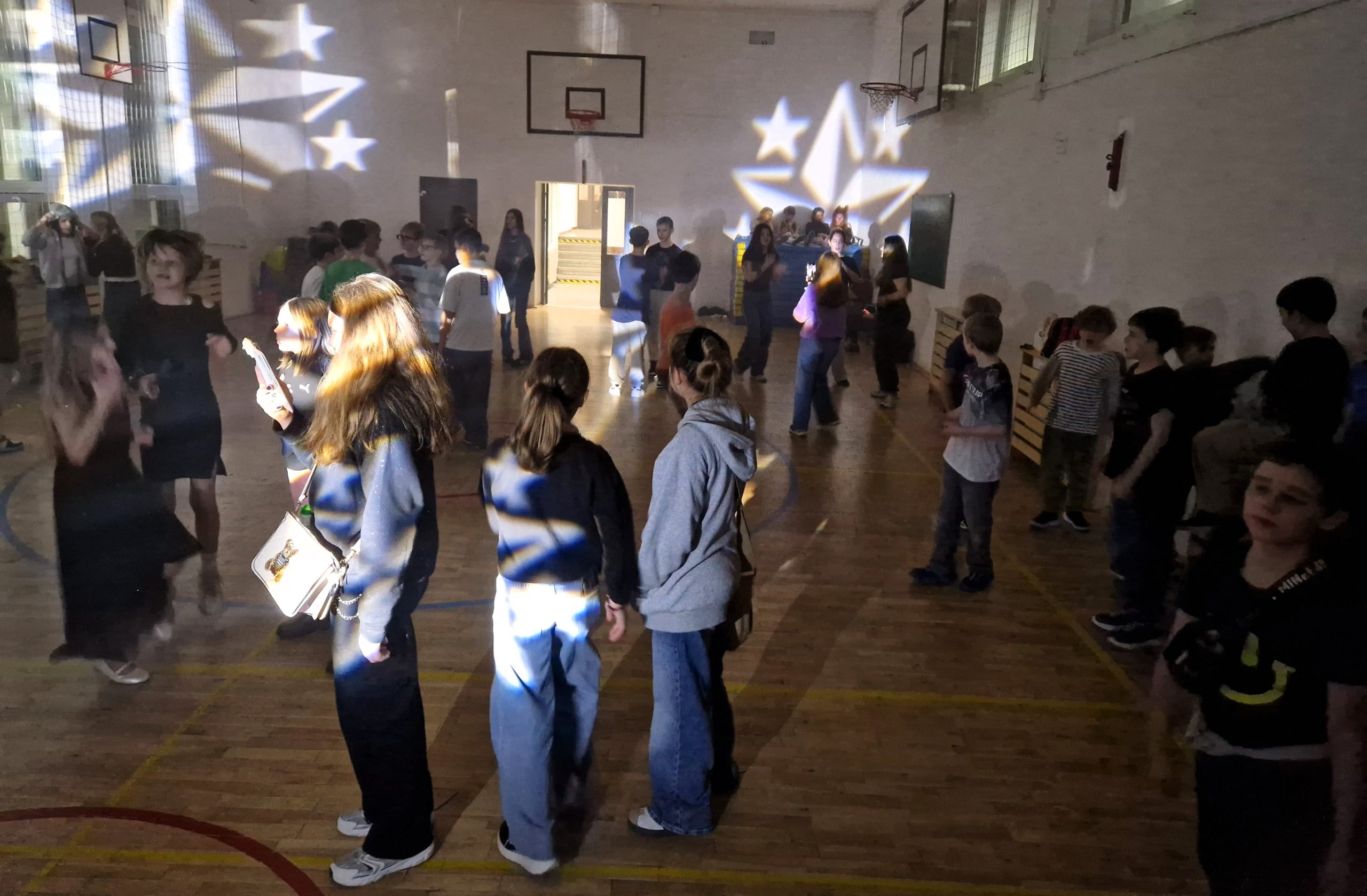 Cztery uczennice stojące razem na sali gimnastycznej podczas dyskoteki andrzejkowej, pozujące do zdjęcia w kolorowym świetle.