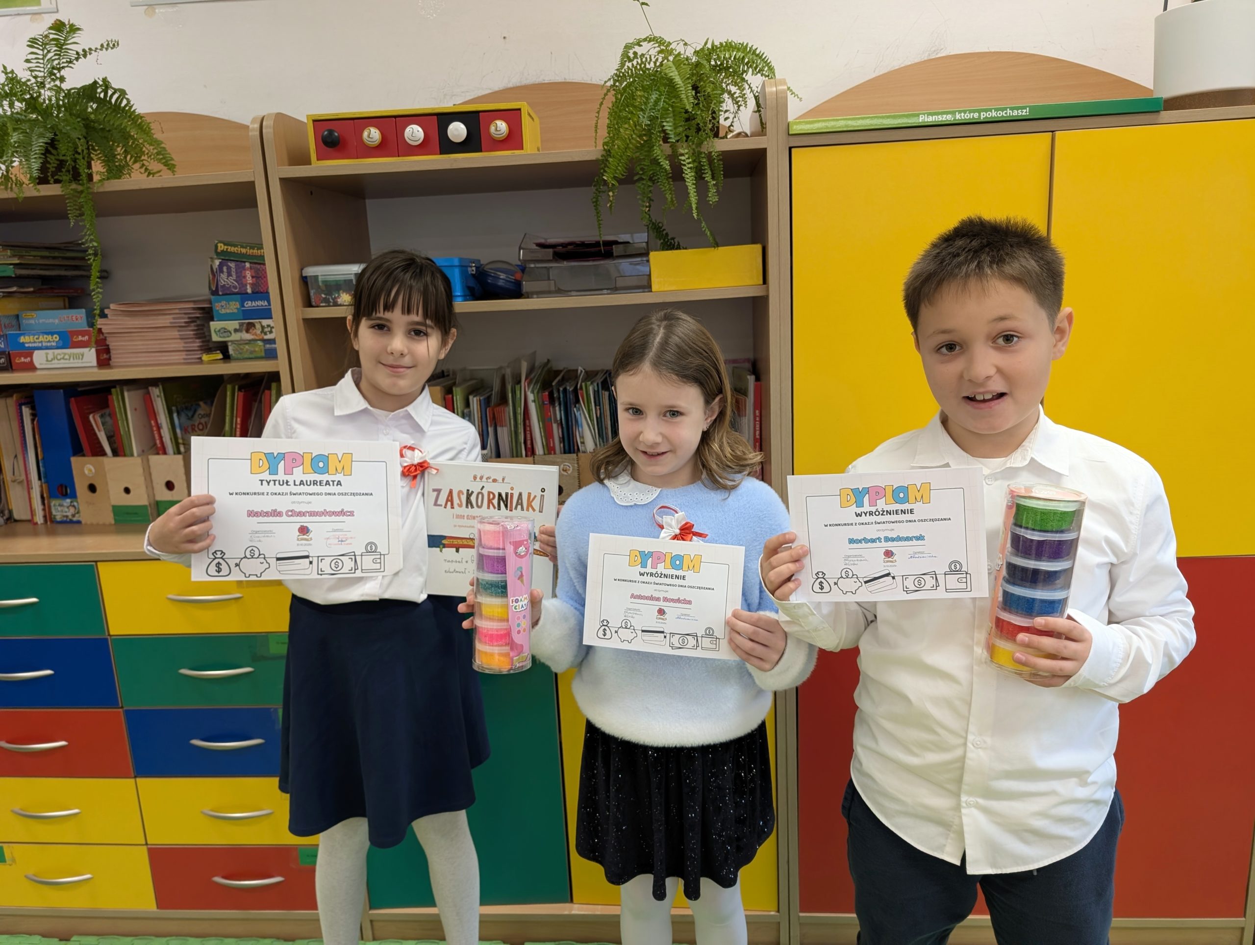 2 uczennice i uczeń z klasy 2a. Trzymają w rękach dyplomy. Dziewczynka po lewej dodatkowo trzyma książkę, a pozostali masę plastyczną w pudełkach. W tle szkolne szafki i szuflady.