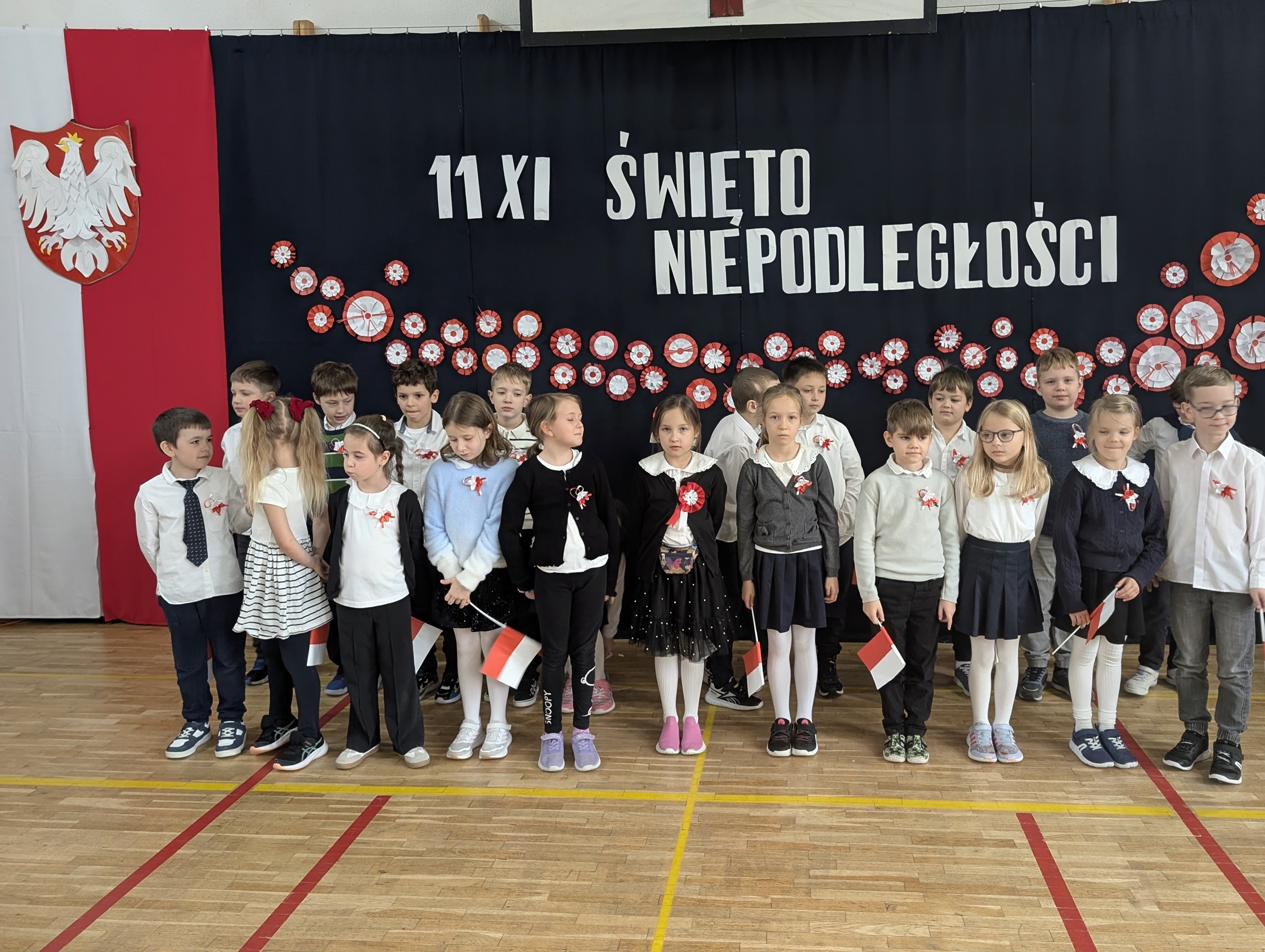 Uczniowie z klasy 2a stoją na sali gimnastycznej przed dekoracją z napisem „11 XI Święto Niepodległości”. Dzieci ubrane są odświętnie, mają przyczepione do ubrań kokardy narodowe, a w rękach trzymają flagi.