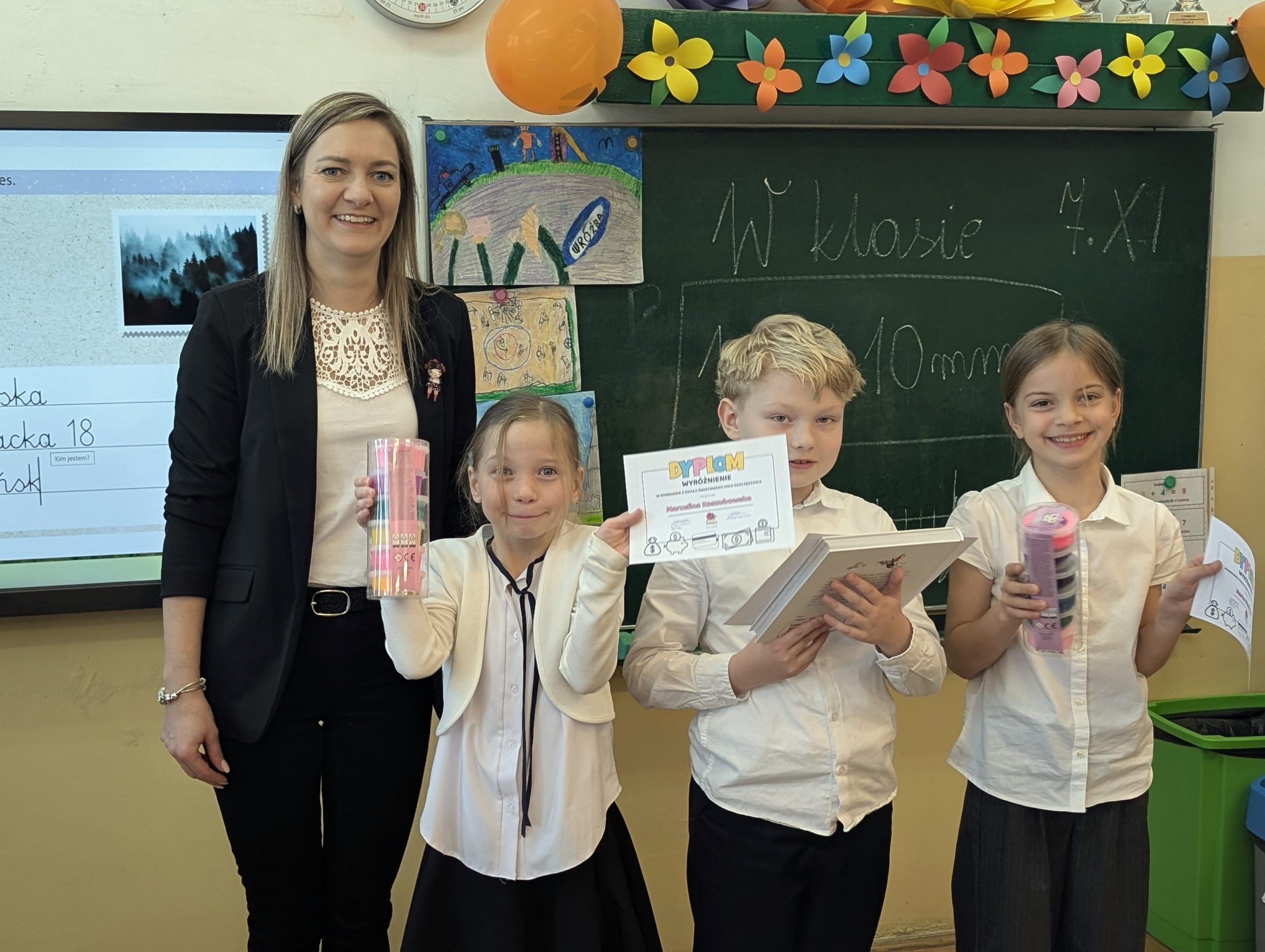 Od lewej vice dyrektor szkoły, uczennica, uczeń i uczennica. Wszyscy są uśmiechnięci. Dzieci trzymają w rękach dyplomy, chłopiec w środku dodatkowo książkę, a dziewczynki masę plastyczną w pudełkach. W tle tablica, telewizor i ozdoby kwietne na tablicą.