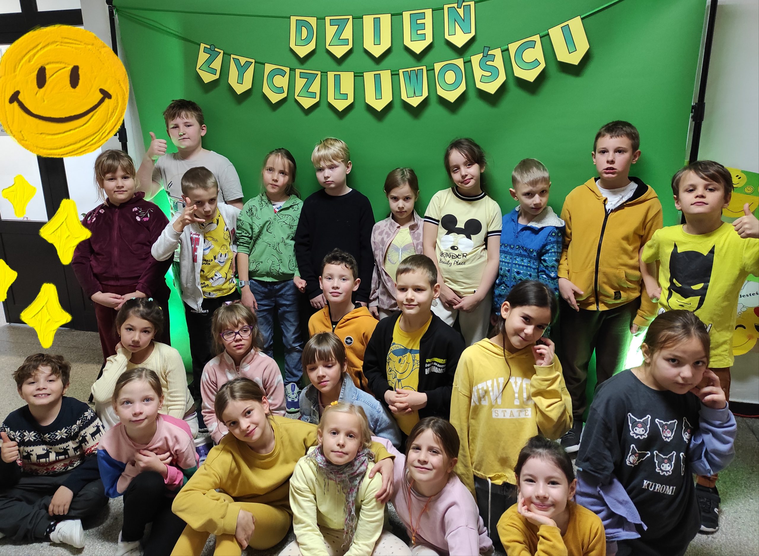 Grupa dzieci ubranych na żółto pozuje z papierowymi emotikonami, balonami i akcesoriami do zdjęć z okazji „Dnia Życzliwości”, stojąc przed zielonym tłem z dekoracyjnym napisem DZIEŃ ŻYCZLIWOŚCI