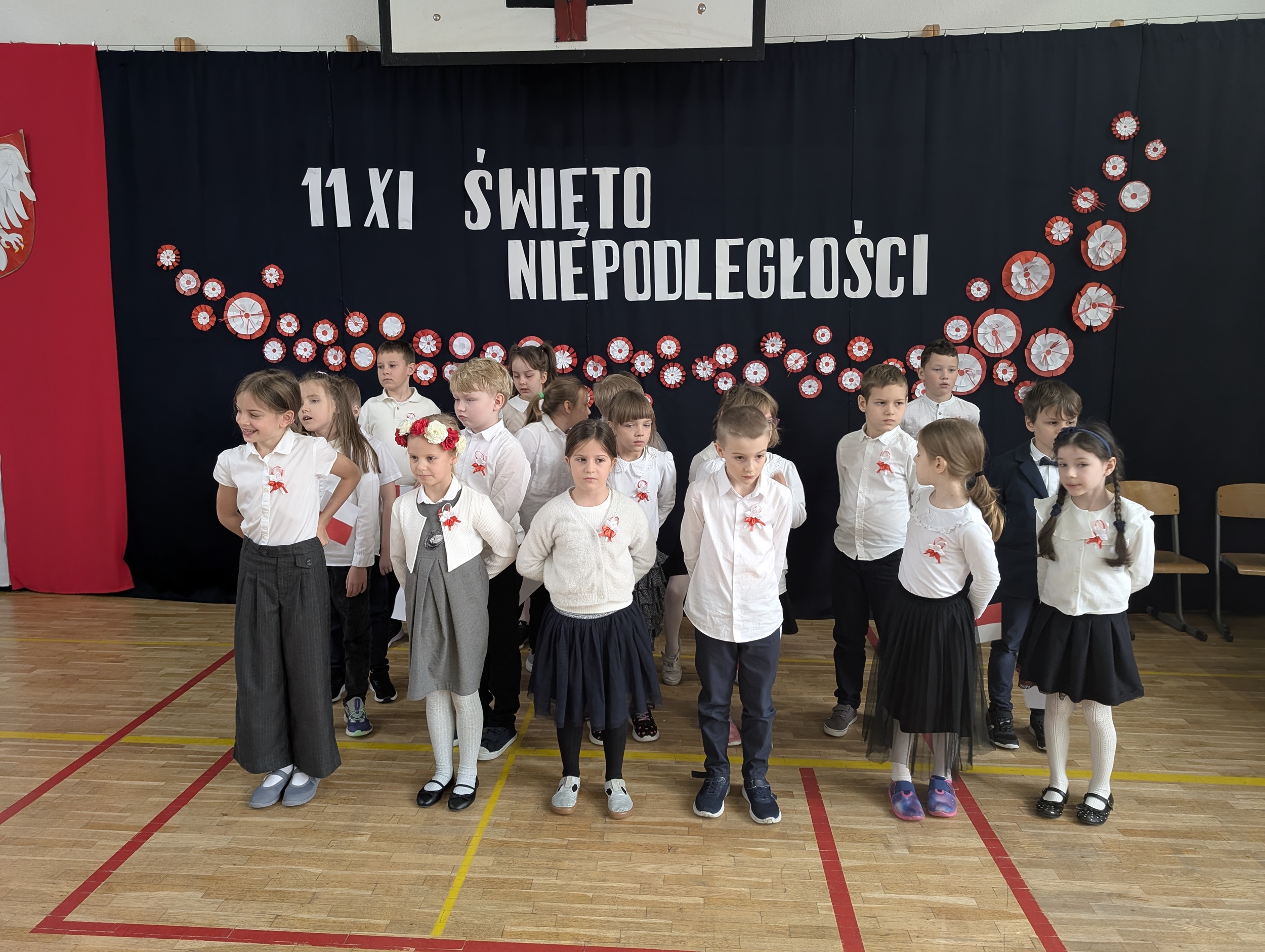 Uczniowie z klasy 2b stoją na sali gimnastycznej przed dekoracją z napisem „11 XI Święto Niepodległości”. Dzieci ubrane są odświętnie, mają przyczepione do ubrań kokardy narodowe.