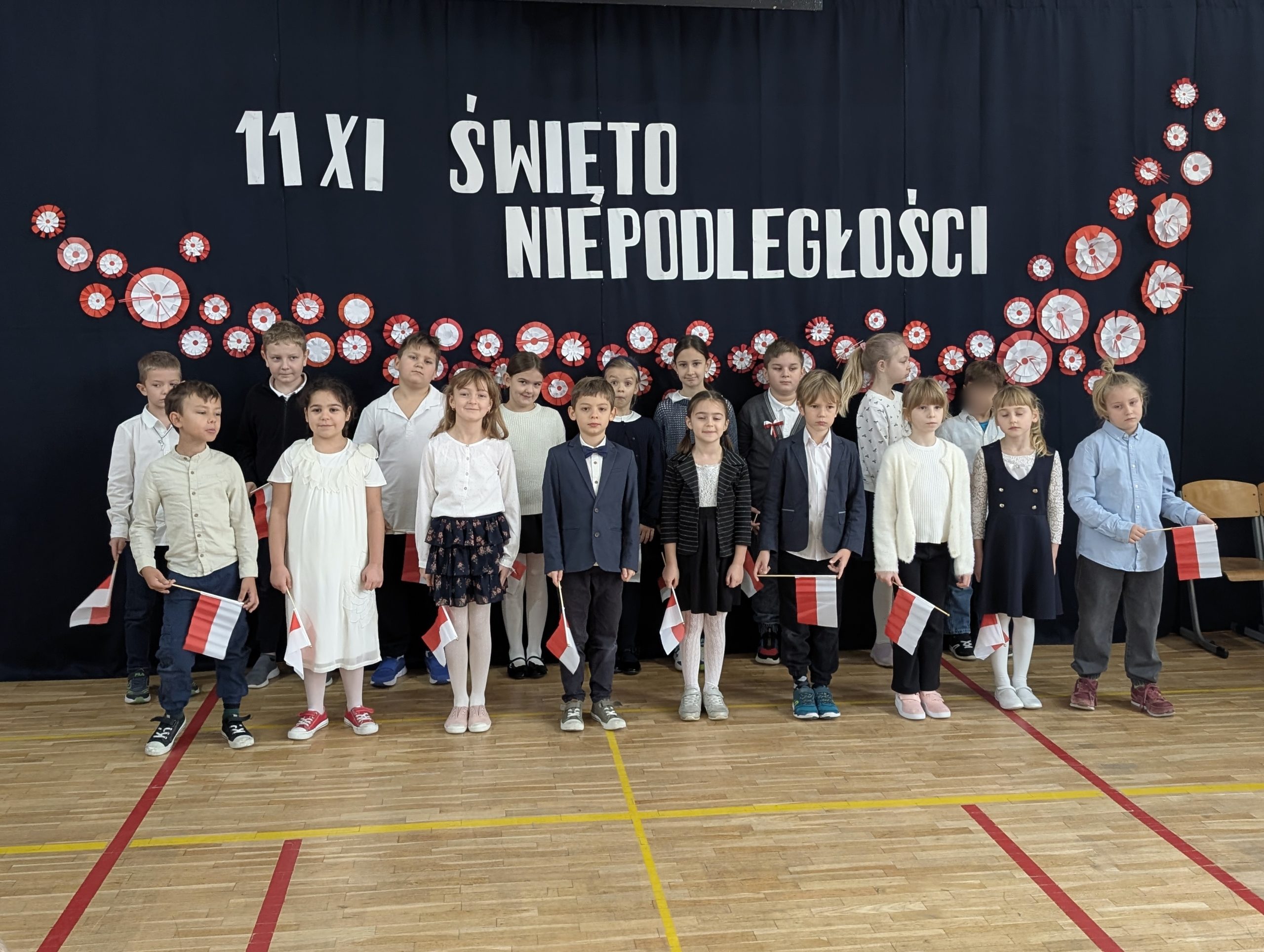 Uczniowie z klasy 2c stoją na sali gimnastycznej przed dekoracją z napisem „11 XI Święto Niepodległości”. Dzieci ubrane są odświętnie, a w dłoniach trzymają flagi.