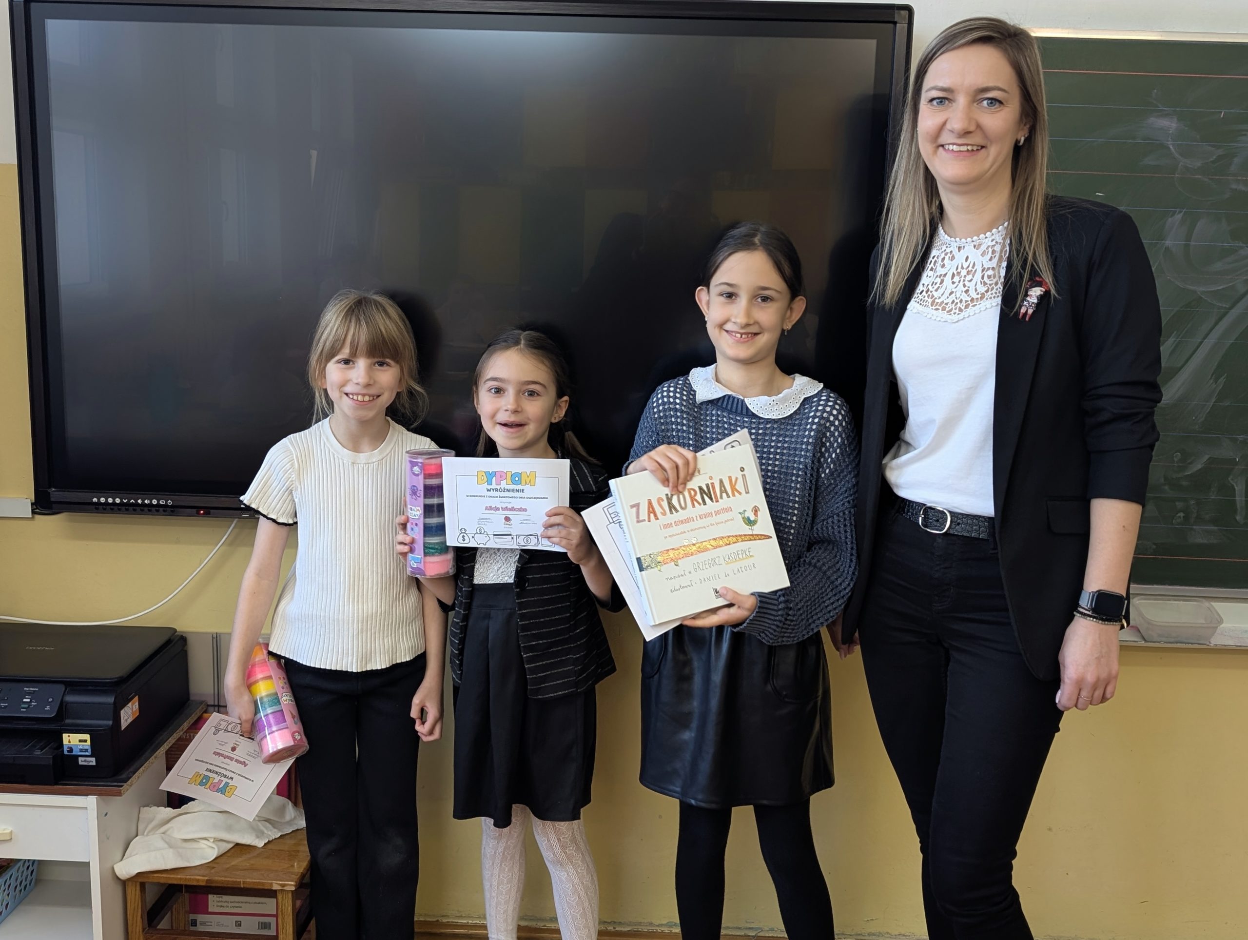 Od prawej stoją vice dyrektor szkoły i 3 uczennice z klasy 2c. Wszyscy są uśmiechnięci. Dzieci trzymają w rękach dyplomy, pierwsza dziewczynka dodatkowo trzyma książkę, a pozostałe masę plastyczną w pudełkach. W tle tablica i telewizor.