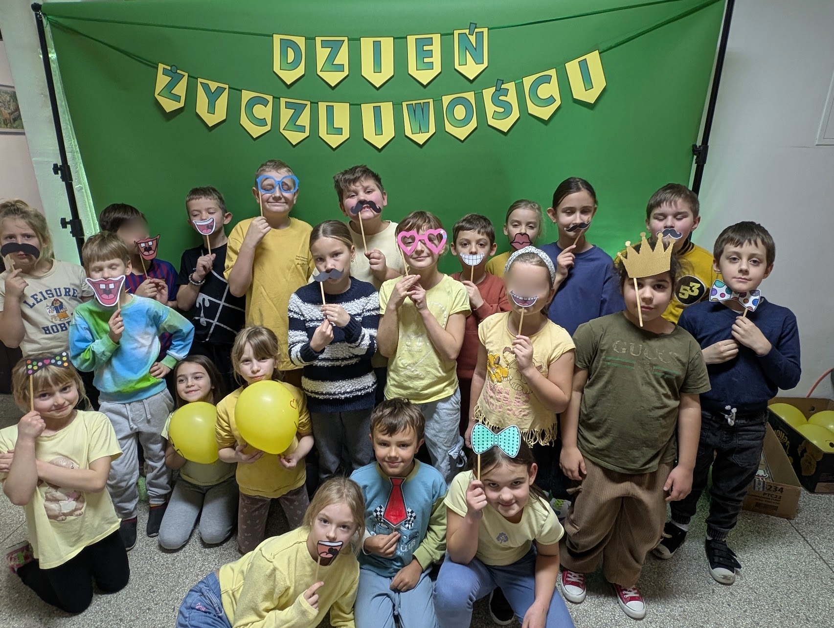 Grupa dzieci ubranych na żółto pozuje z papierowymi emotikonami, balonami i akcesoriami do zdjęć z okazji „Dnia Życzliwości”, stojąc przed zielonym tłem z dekoracyjnym napisem DZIEŃ ŻYCZLIWOŚCI